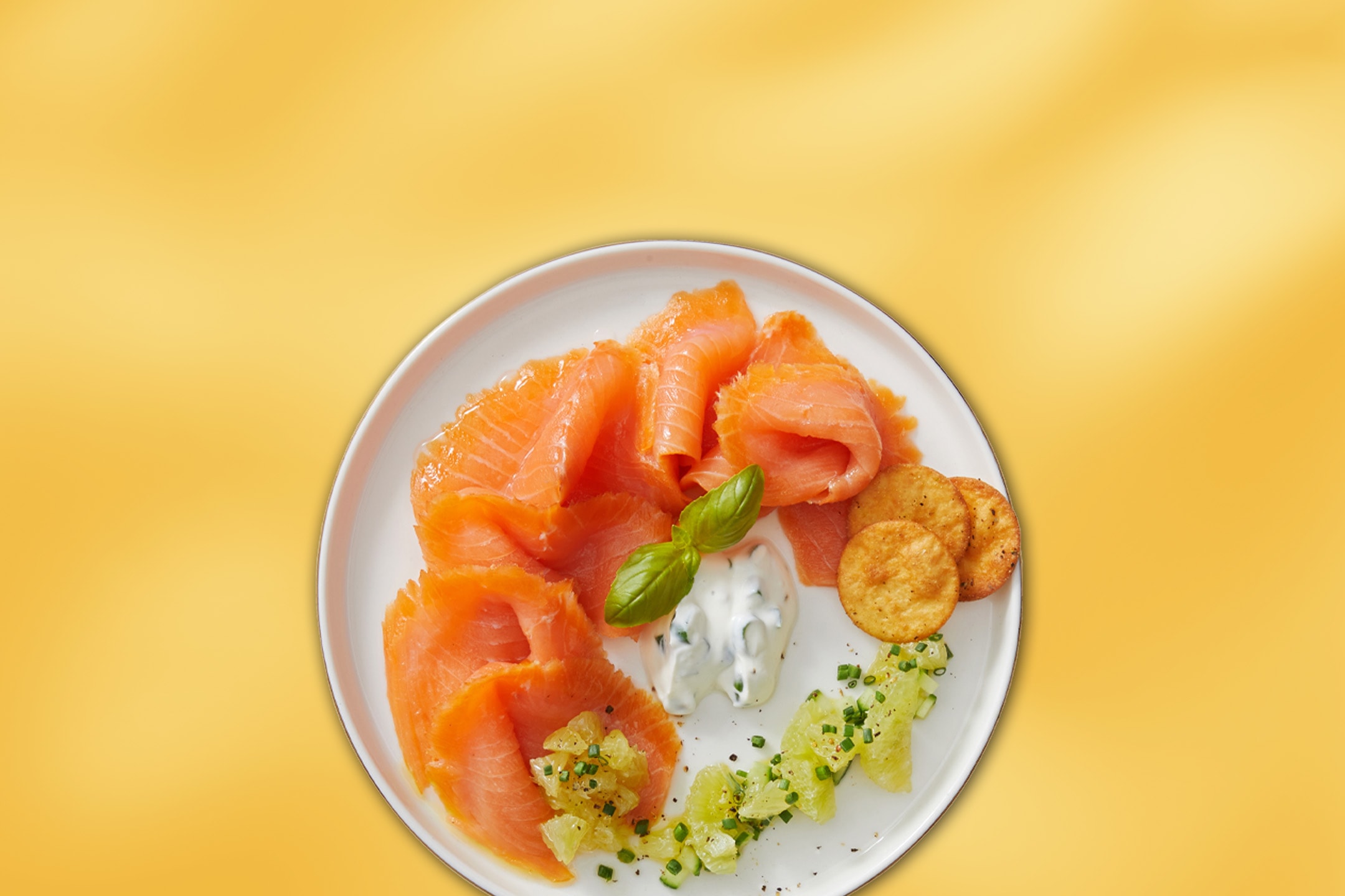 Røget laks med creme fraiche, kiks og basilikum på en hvid tallerken.