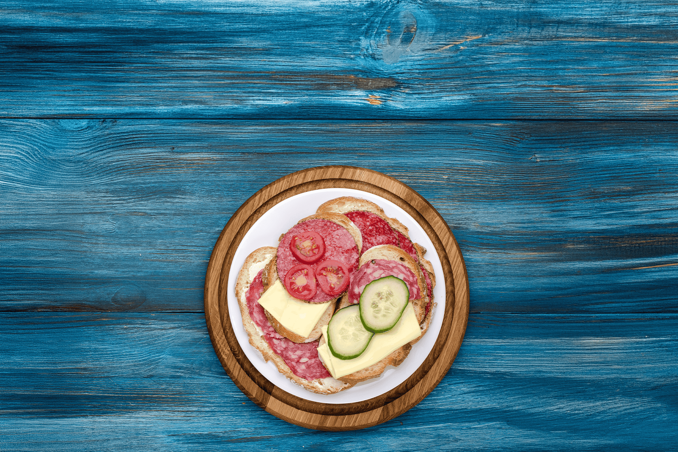 En tallerken med salamimadder med ost, agurk og tomat på et blåt træbord.
