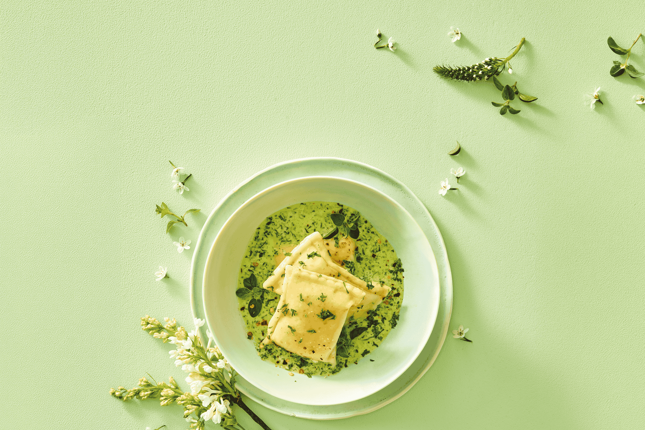 Ravioli i grøn sauce med krydderurter og blomster på en lysegrøn baggrund.