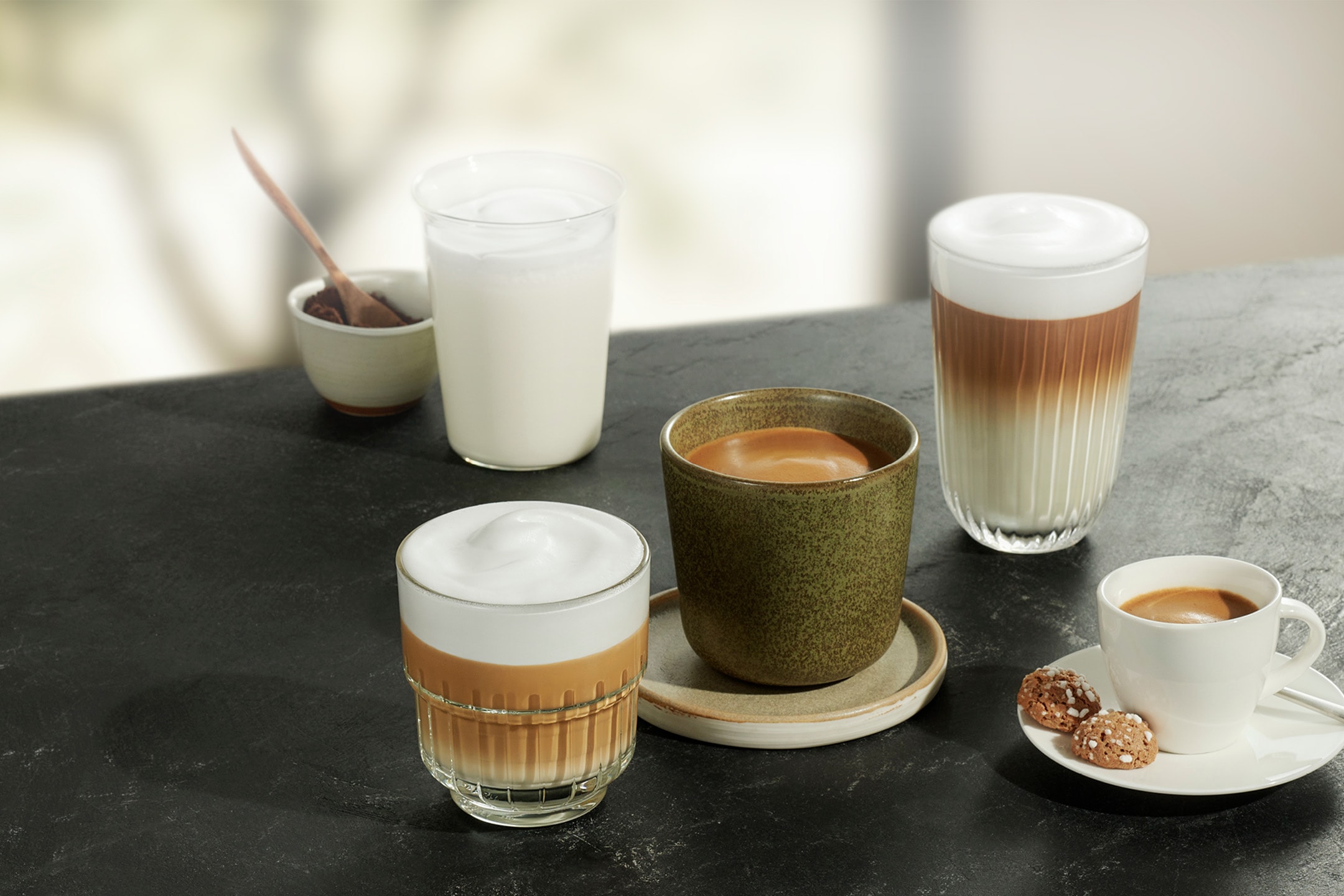 Forskellige kaffedrikke, herunder latte, cappuccino og espresso, på et mørkt bord.