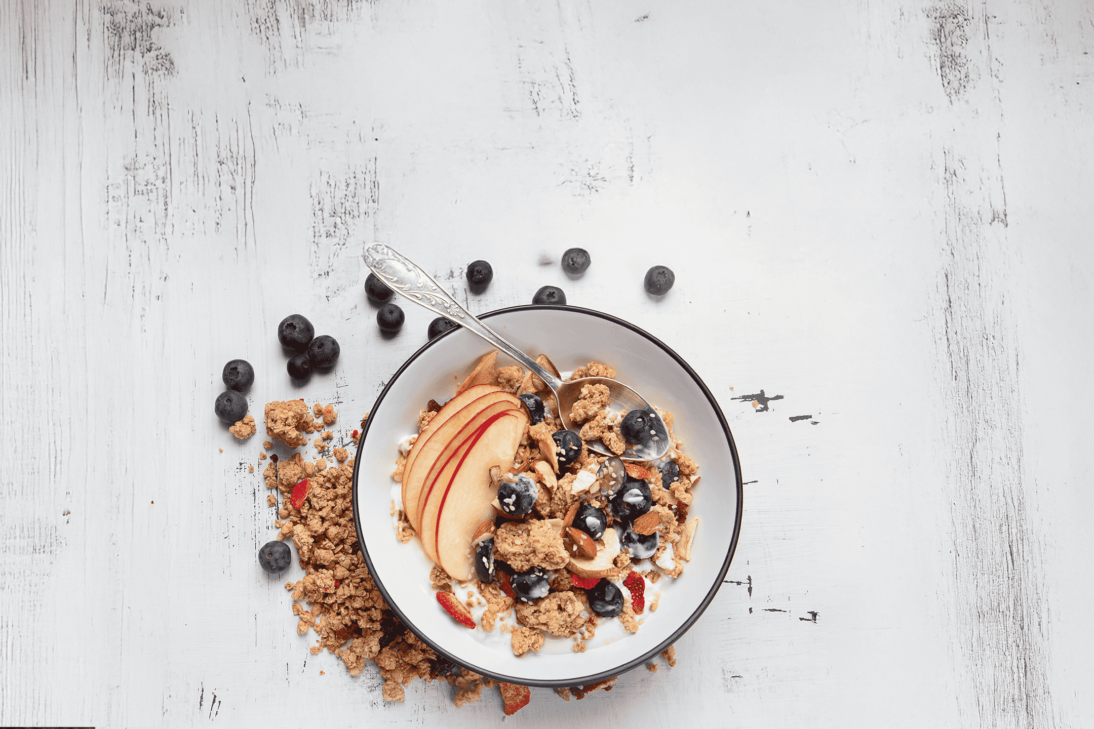 Morgenmadsskål med granola, yoghurt, blåbær, æbleskiver og nødder på et hvidt træbord.