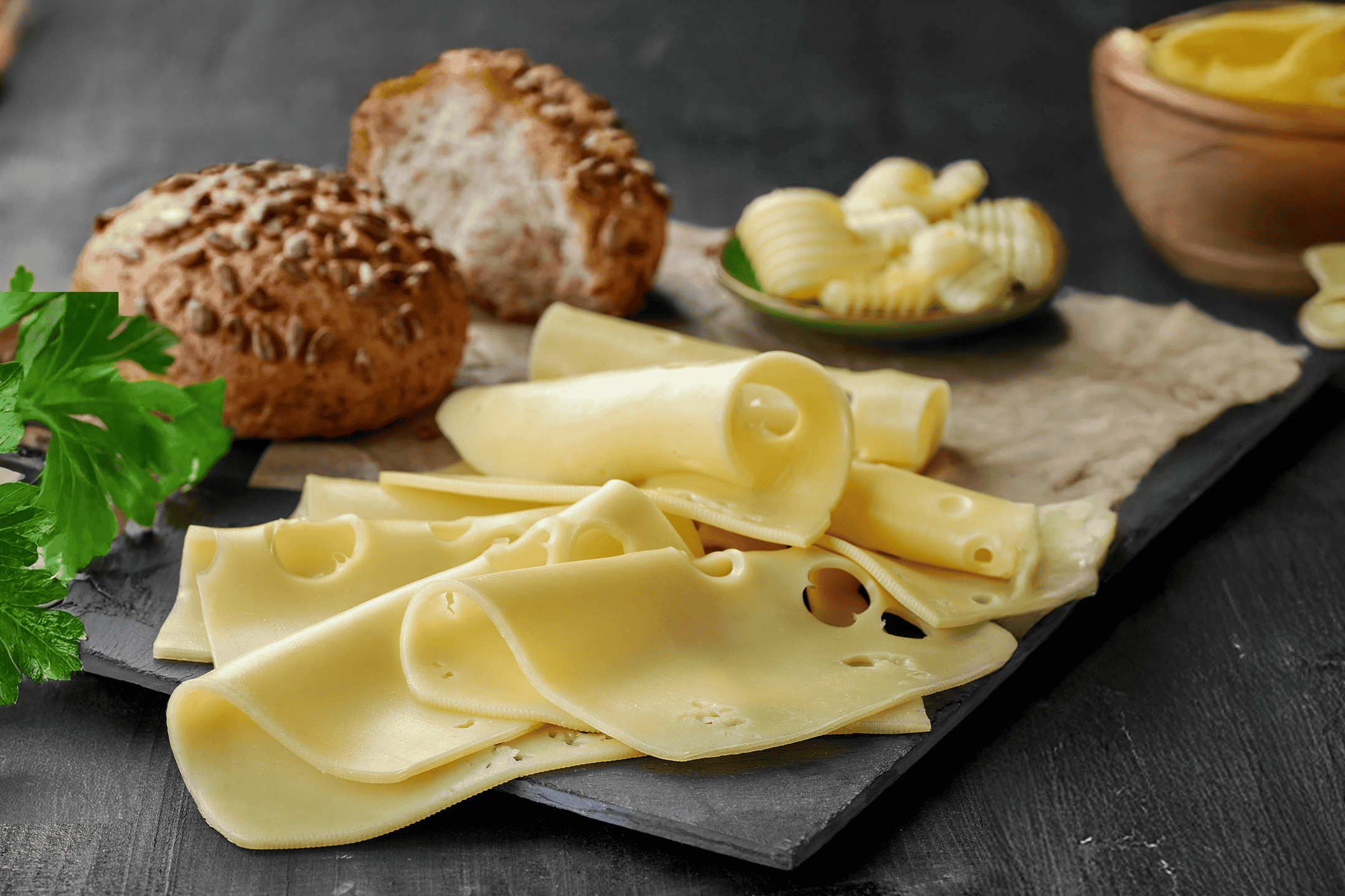 Skiver af ost, smørkrøller og brødboller på en mørk skiferplade.