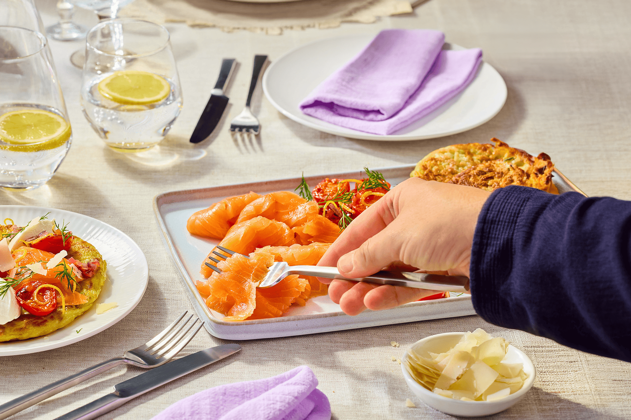 En person spiser røget laks og kartoffelpandekager ved et bord dækket med lilla servietter og citronvand.