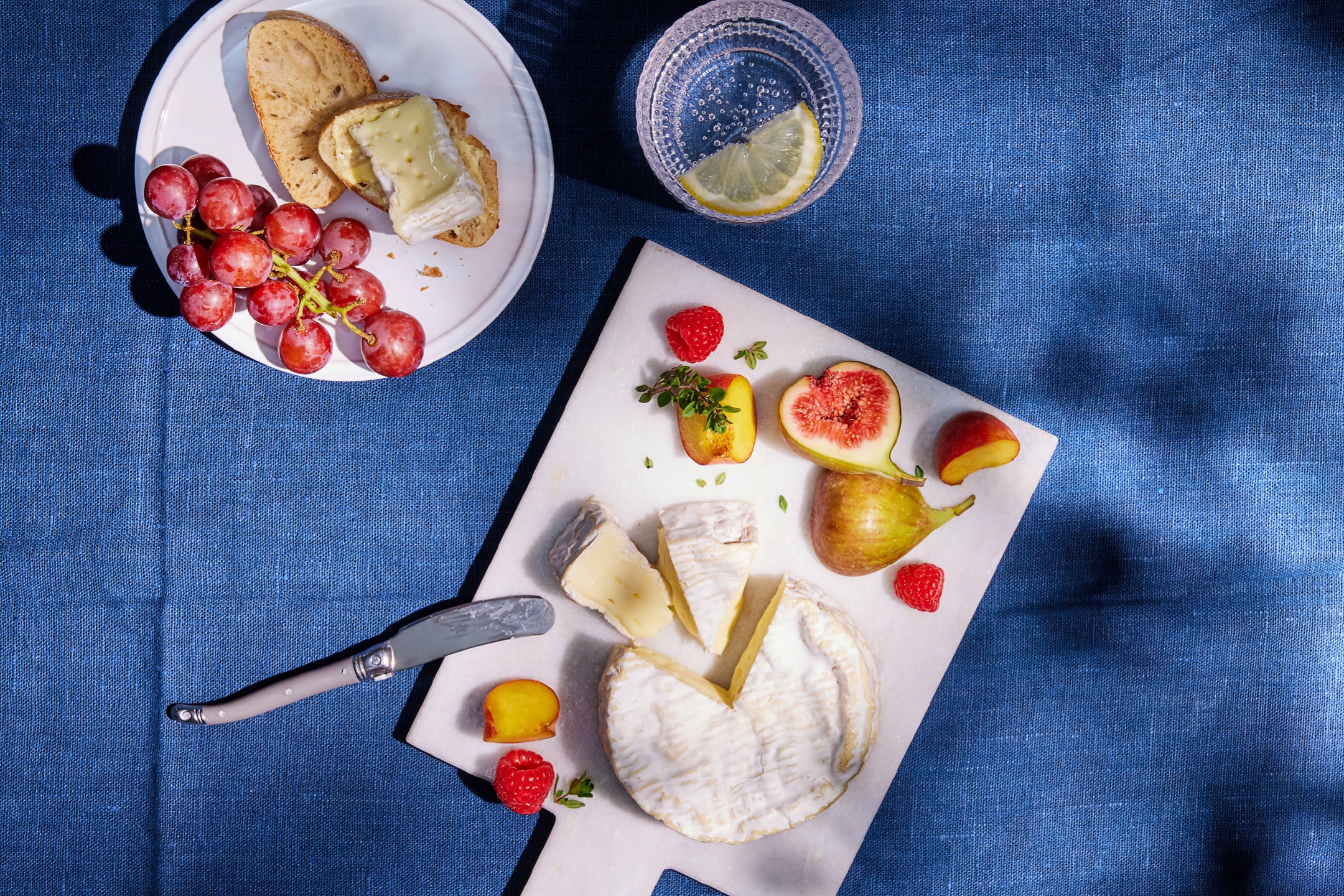 En tallerken med brieost, brød, røde druer, figner, hindbær og et glas vand med citron på et blåt klæde.
