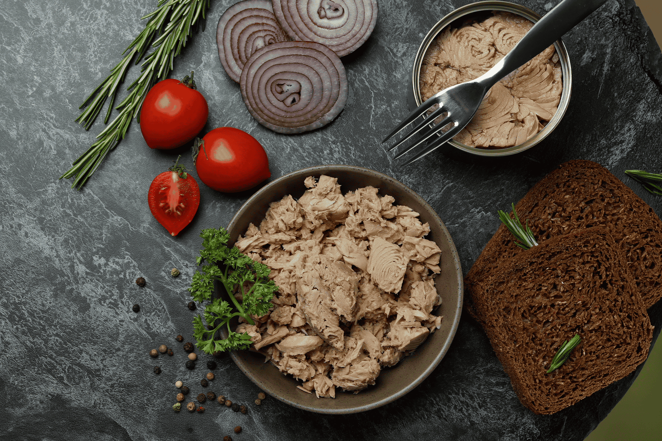 Tun i skål og dåse med brød, tomater, løg og rosmarin på mørk baggrund.