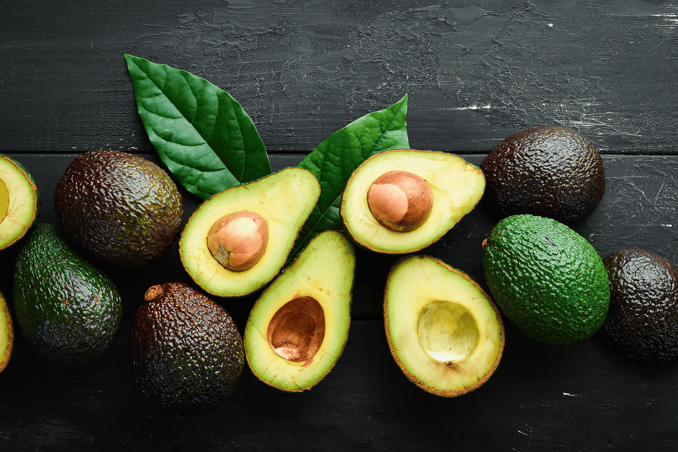 Friske hele og halverede avocadoer med blade på en mørk baggrund.