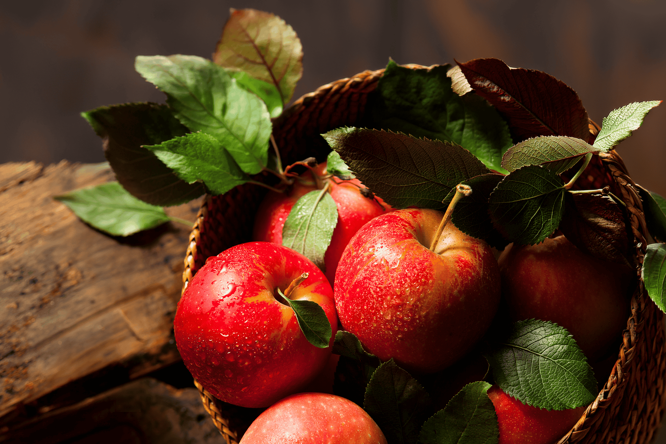 Røde æbler med grønne blade i en flettet kurv på et træbord.