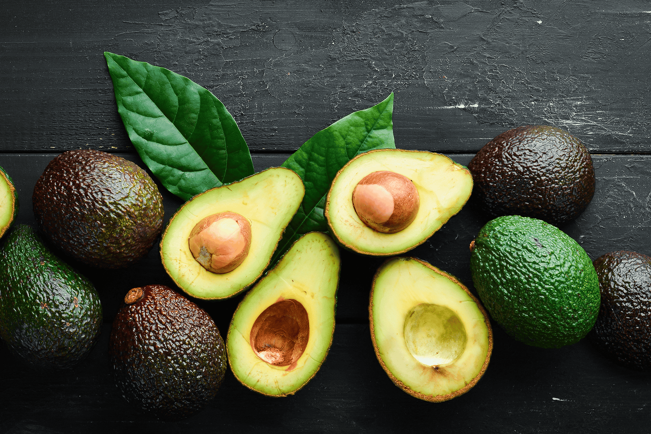 Forskellige modne avocadoer, hele og halverede, med blade på en mørk baggrund.