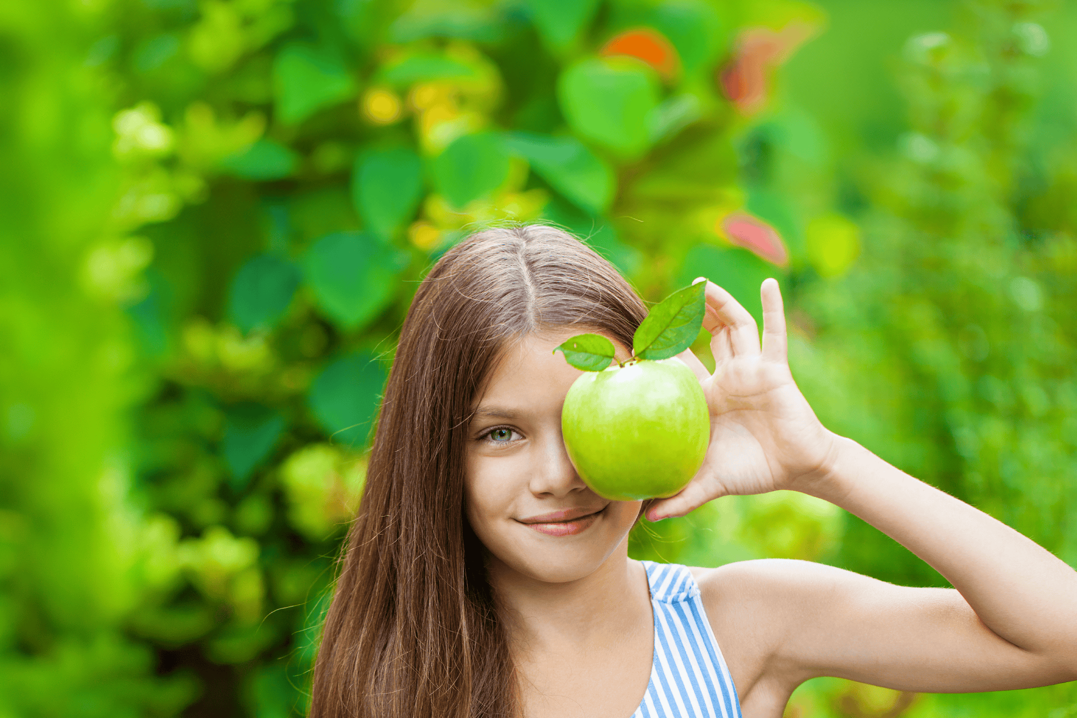 Pige holder et grønt æble foran sit ansigt i en grøn have.