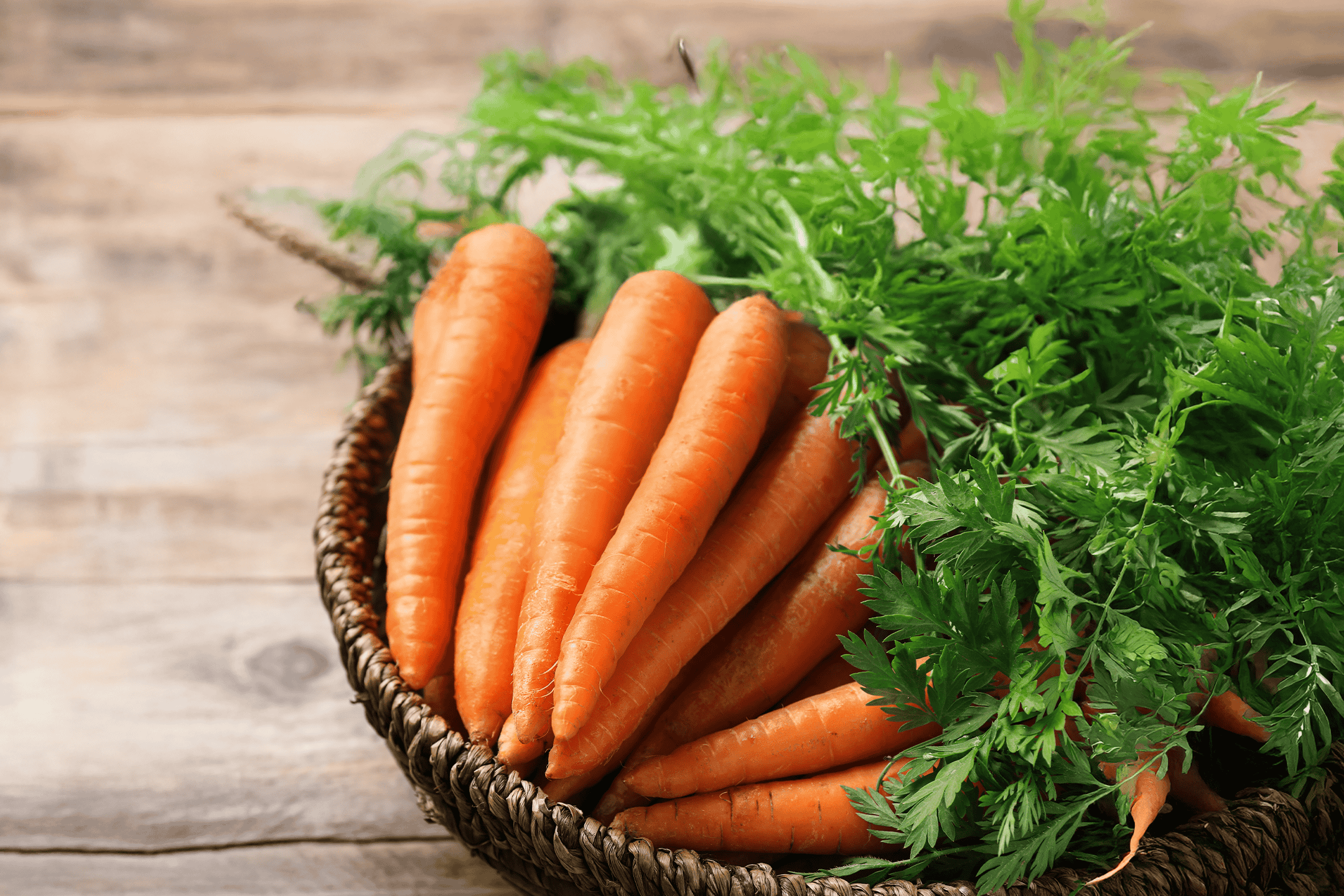 Friske gulerødder med grøn top i en flettet kurv på et træbord.