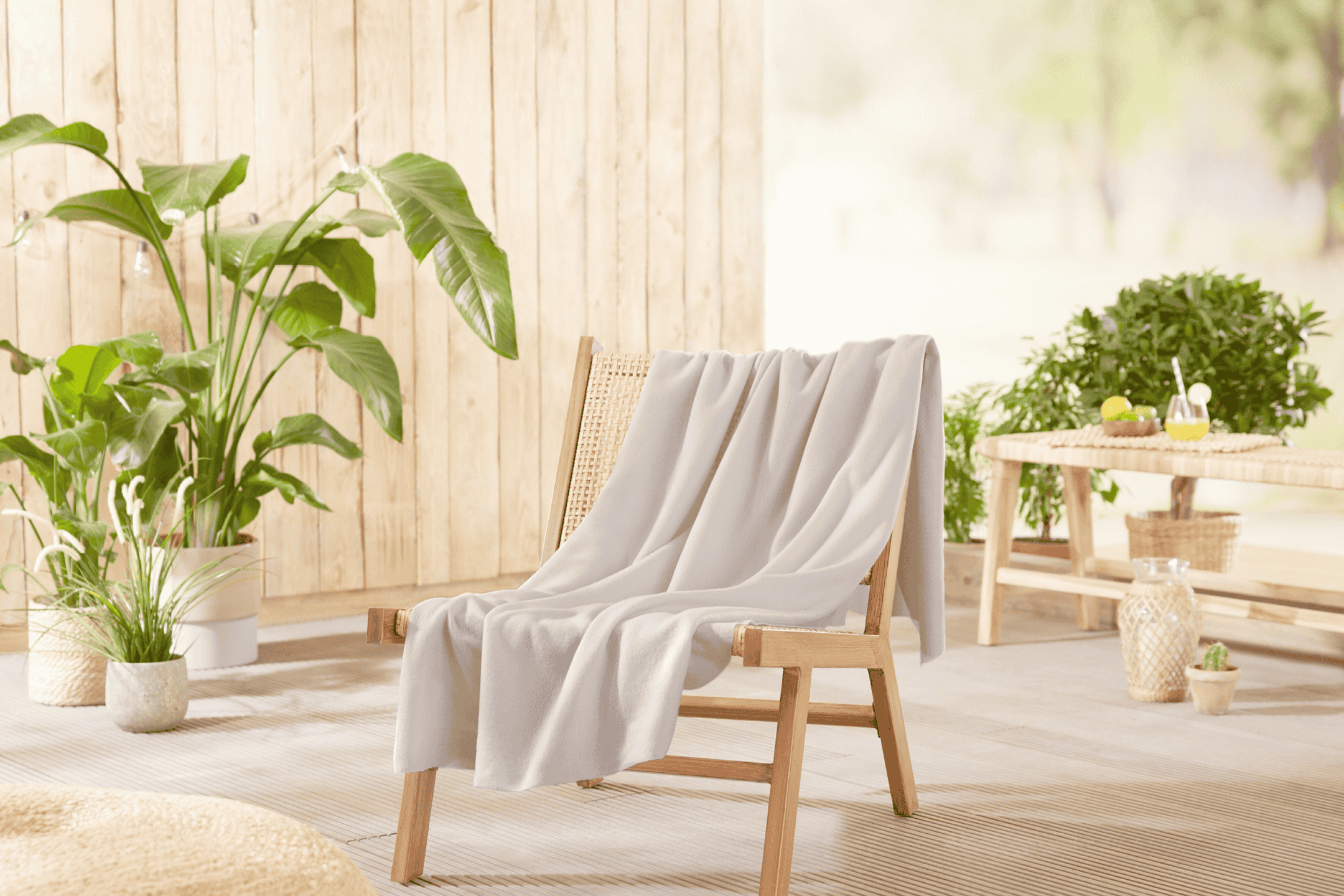 Træstol med lyst tæppe på en terrasse med planter og et bord med drikkevarer.