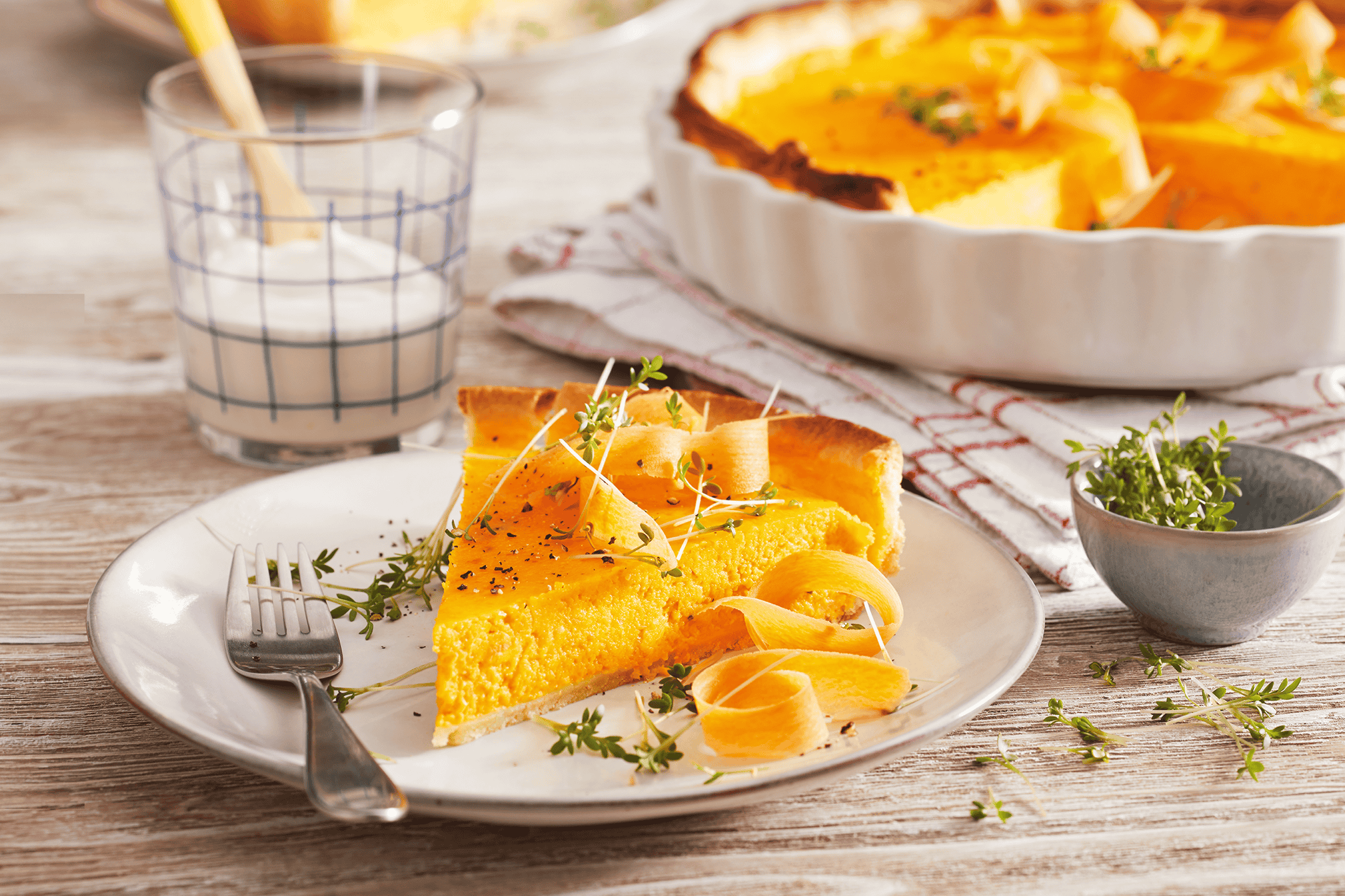 Et stykke græskartærte med gulerodsstrimler og karse på en tallerken, med en hel tærte i baggrunden.