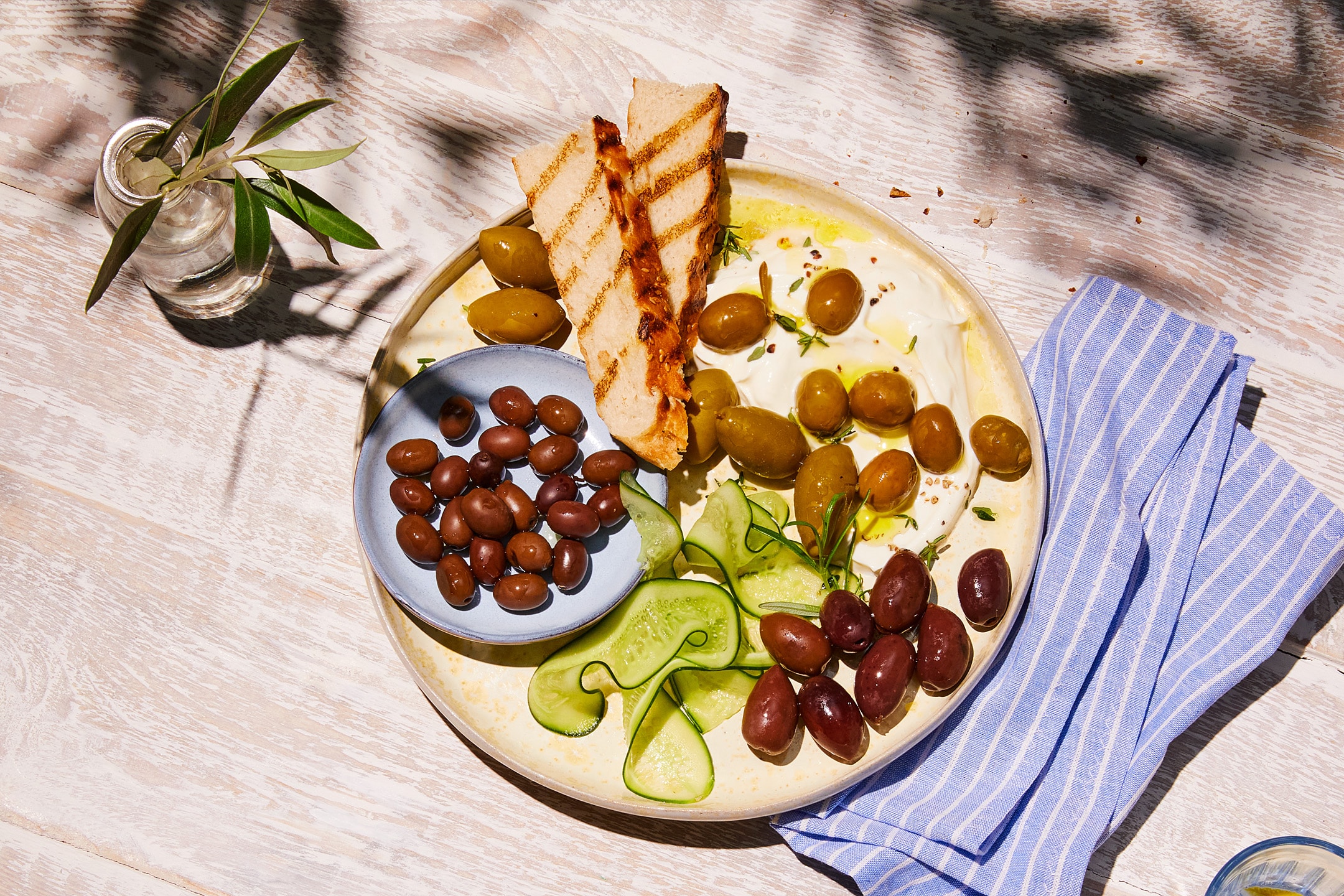 En tallerken med græsk yoghurt, oliven, grillet brød og agurkeskiver på et træbord.