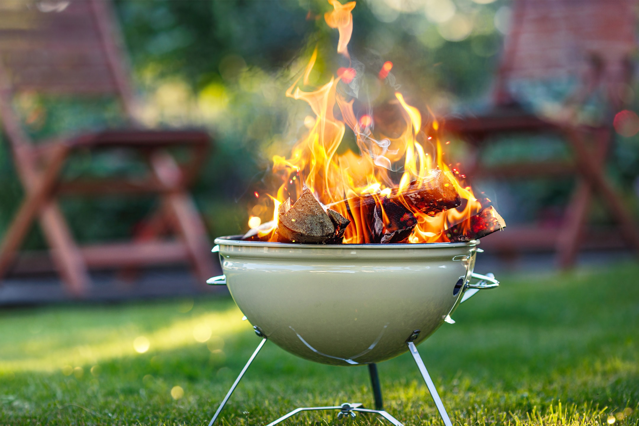 Grill med brændende trækul og flammer på en græsplæne med slørede stole i baggrunden.
