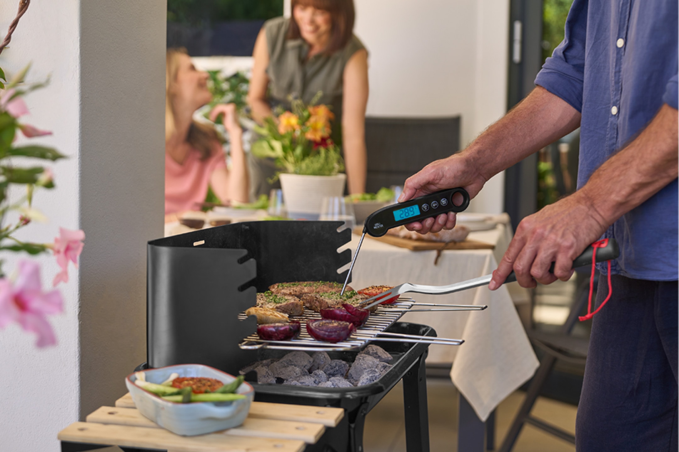 Mand griller kød og grøntsager med et stegetermometer, mens to kvinder hygger sig i baggrunden.