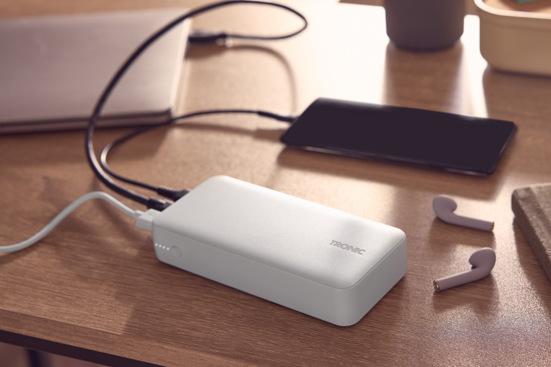 Hvid powerbank oplader en smartphone og en bærbar computer på et træbord, med trådløse høretelefoner.