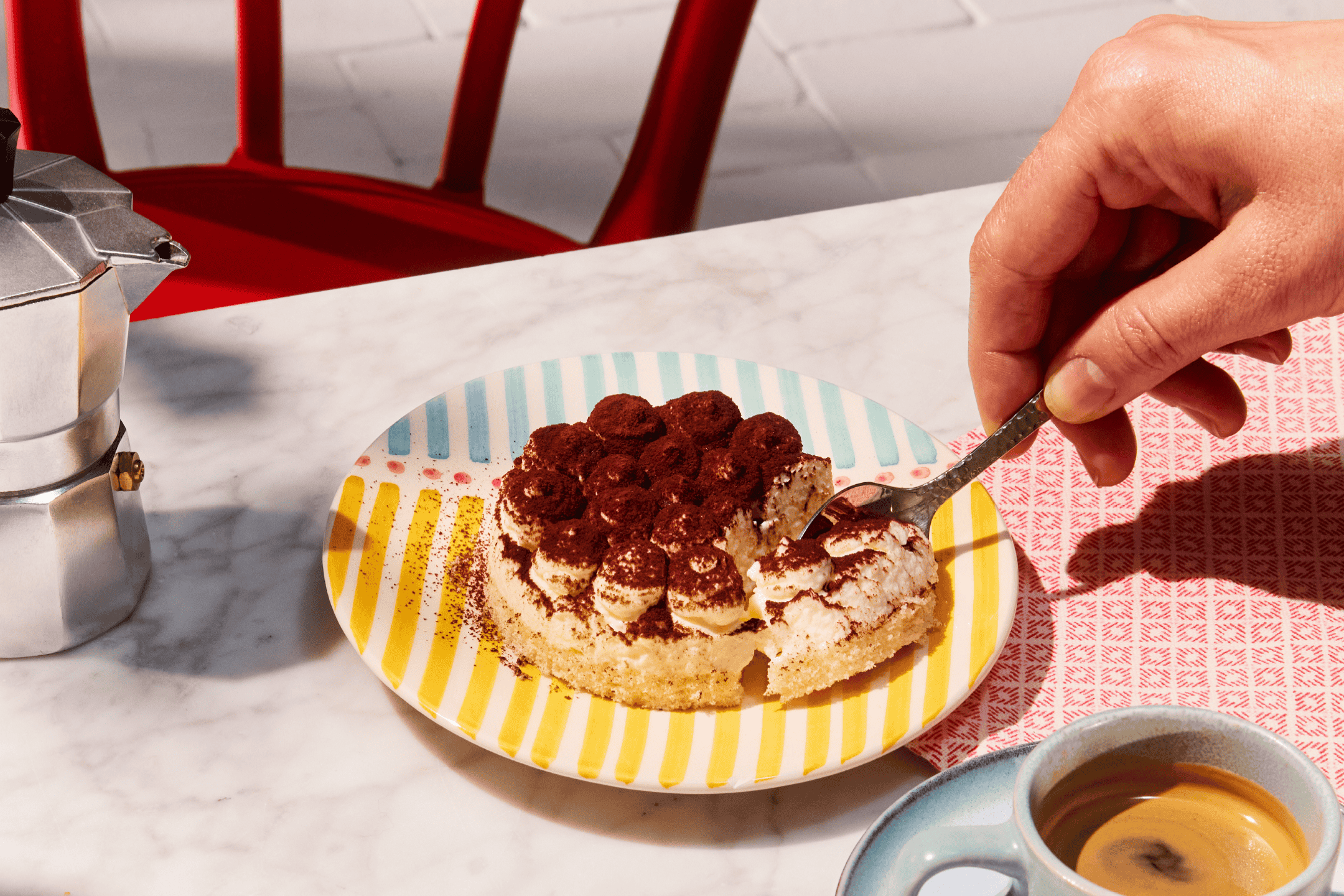 En hånd tager et stykke tiramisu fra en farverig tallerken, med en mokkakande og kaffe.