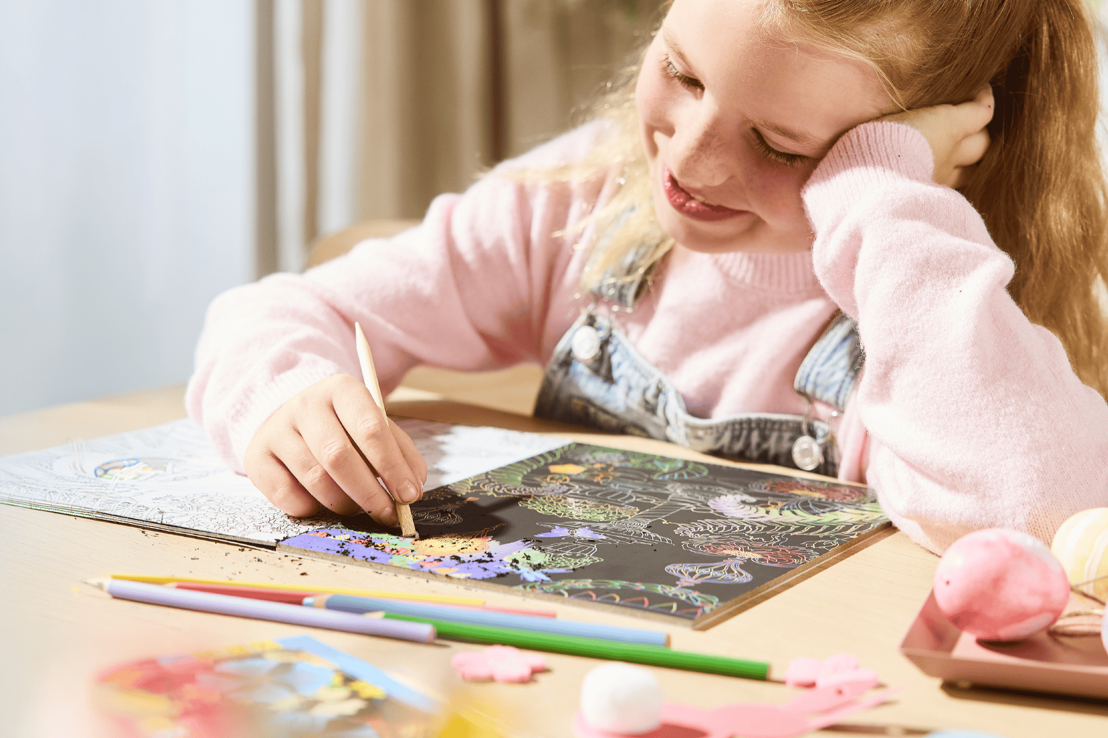 Glad pige laver kradsekunst med farveblyanter på et bord.