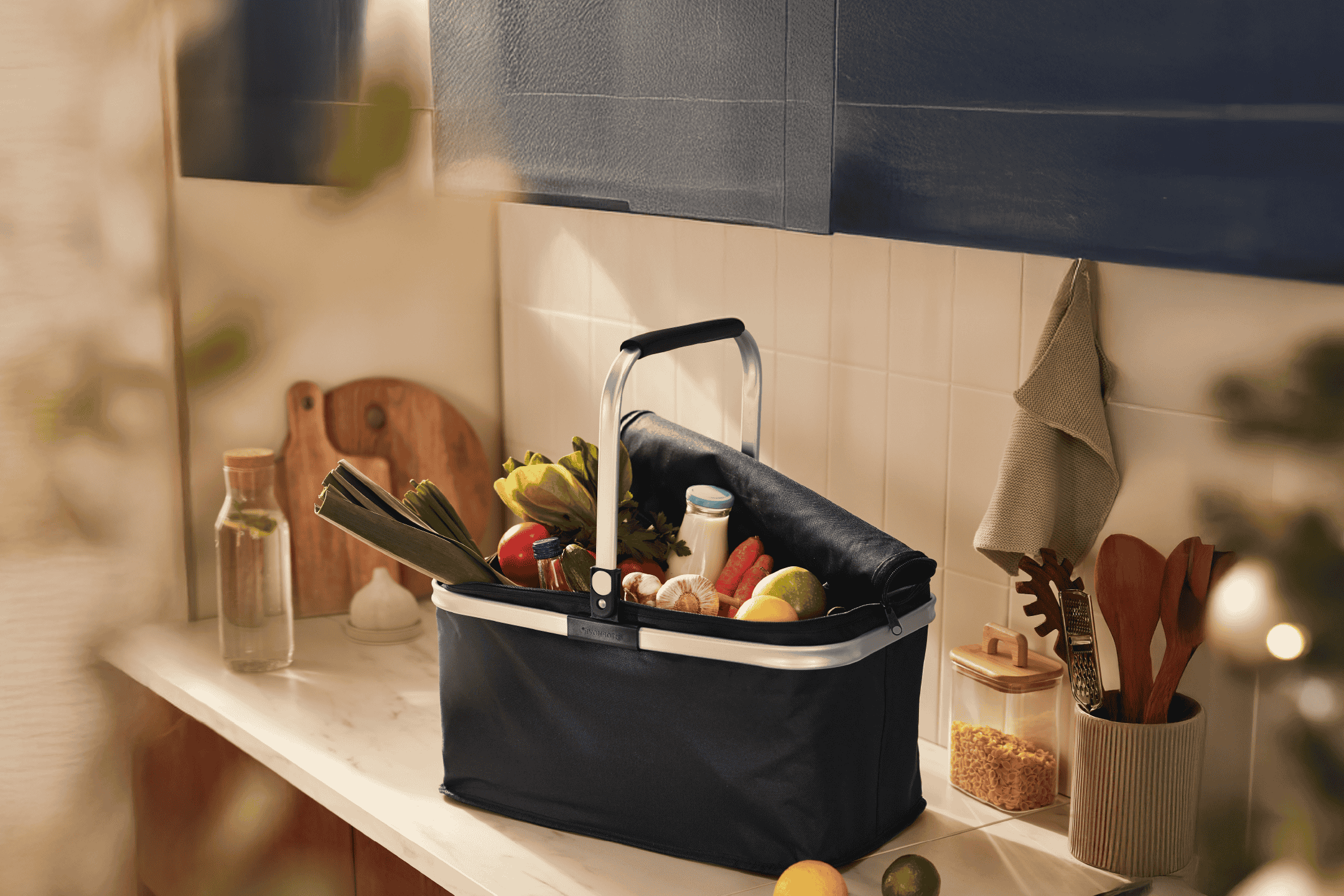 Indkøbskurv fyldt med friske grøntsager og dagligvarer på et køkkenbord.