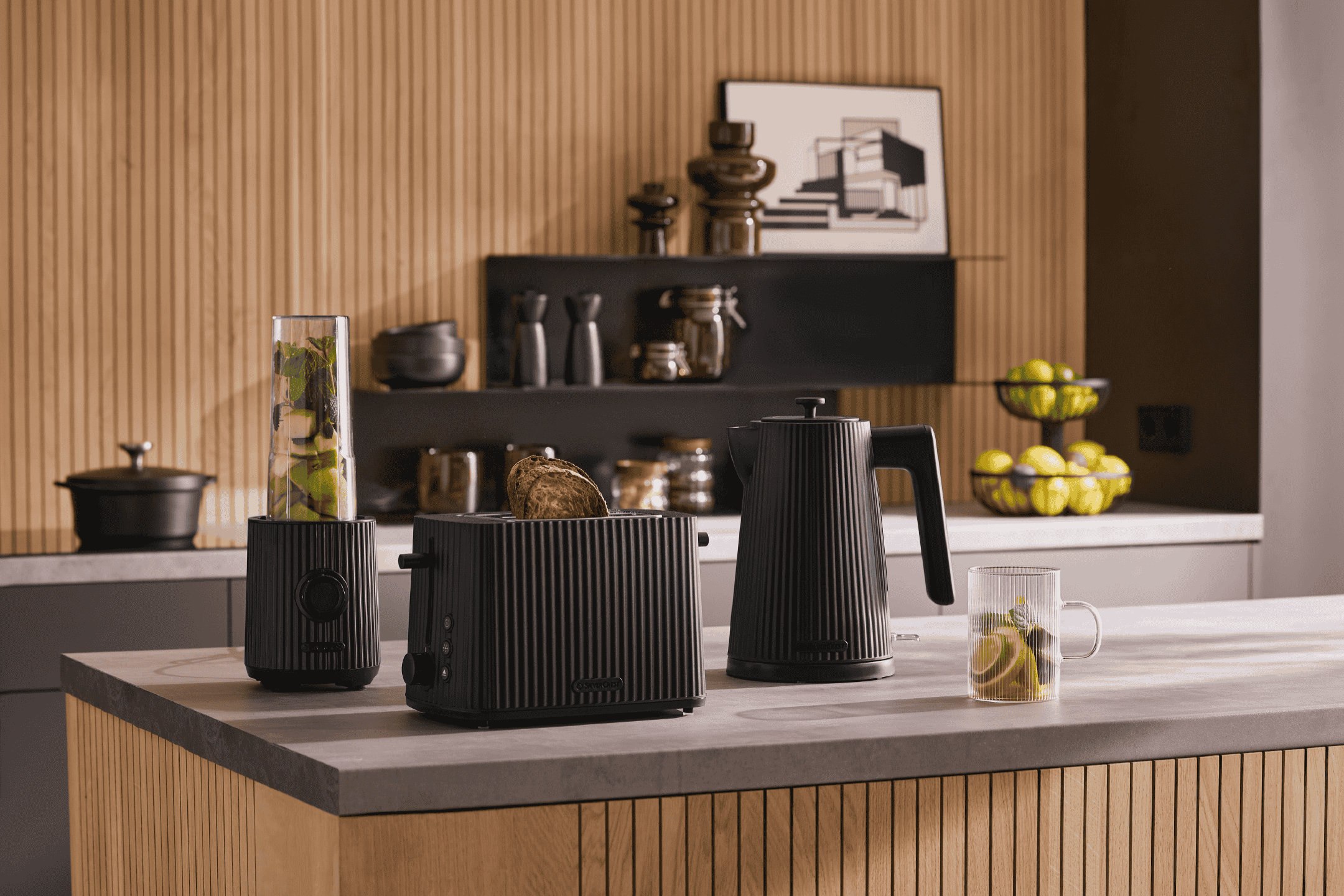 Køkkenbord med sort blender, brødrister med toast og elkedel, samt en kop med lime og mynte.