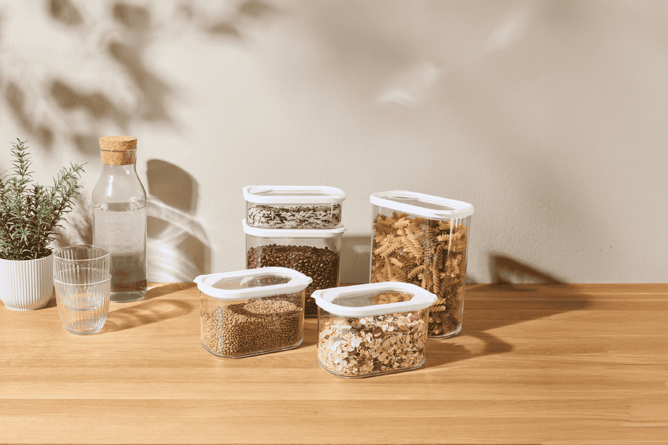 Gennemsigtige opbevaringsbeholdere med tørvarer som pasta, linser og kaffe på et træbord.