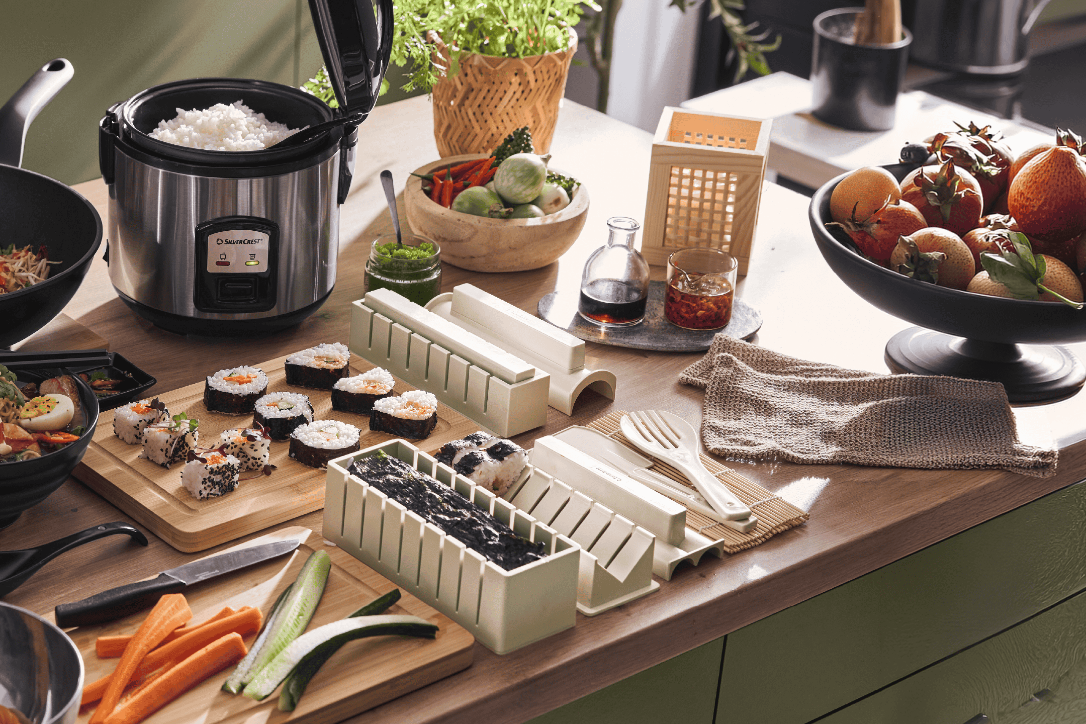Riskoger med ris, sushisæt, skærebræt med grøntsager og frugtskål på et køkkenbord.