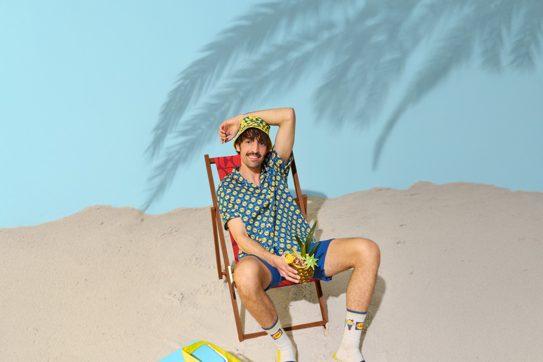 Mand i strandstol med ananas, iført sommerhat, skjorte, shorts og sokker med mønster.