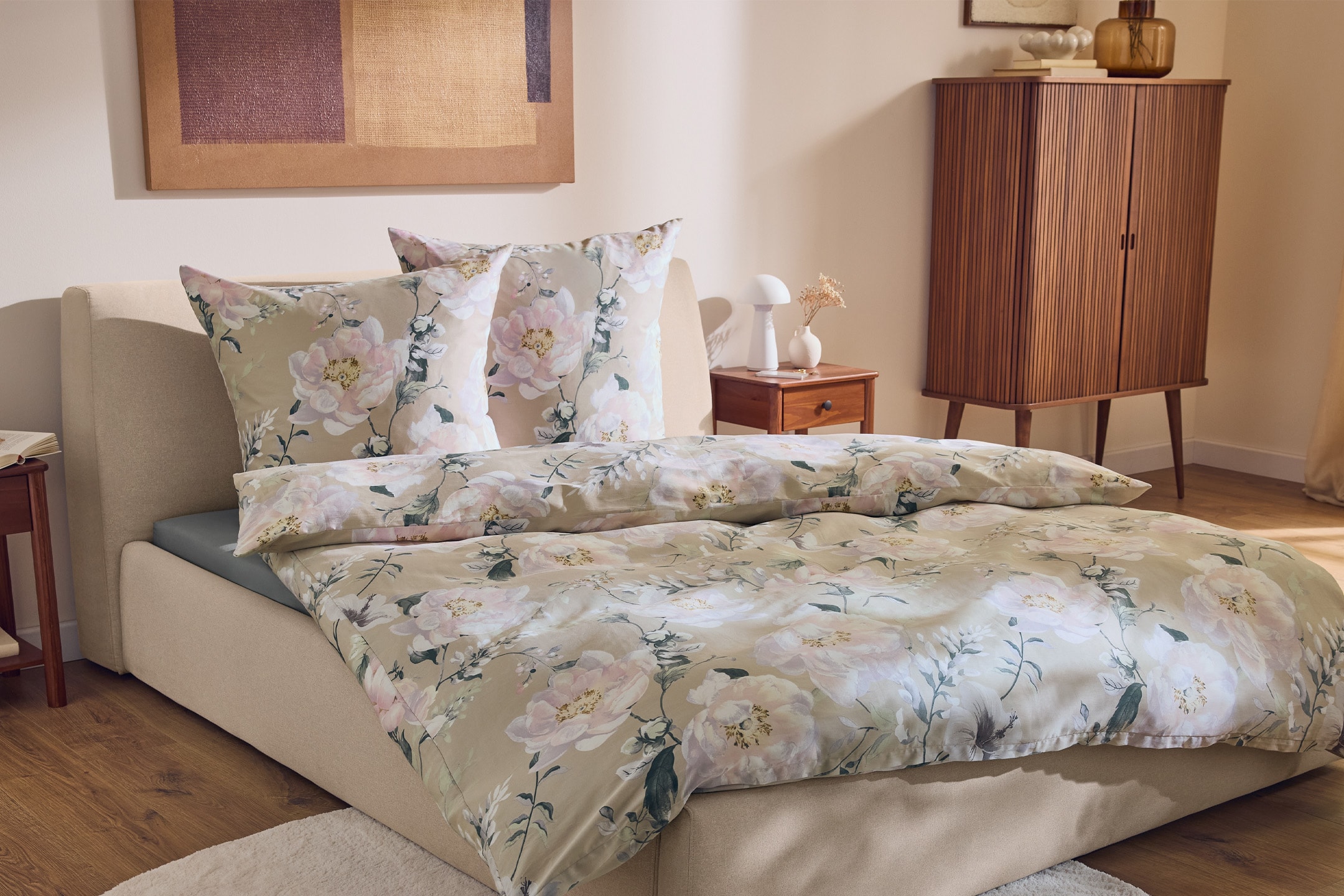 Senge med blomstret sengetøj i beige og lyserøde nuancer, et sengebord og et skab.