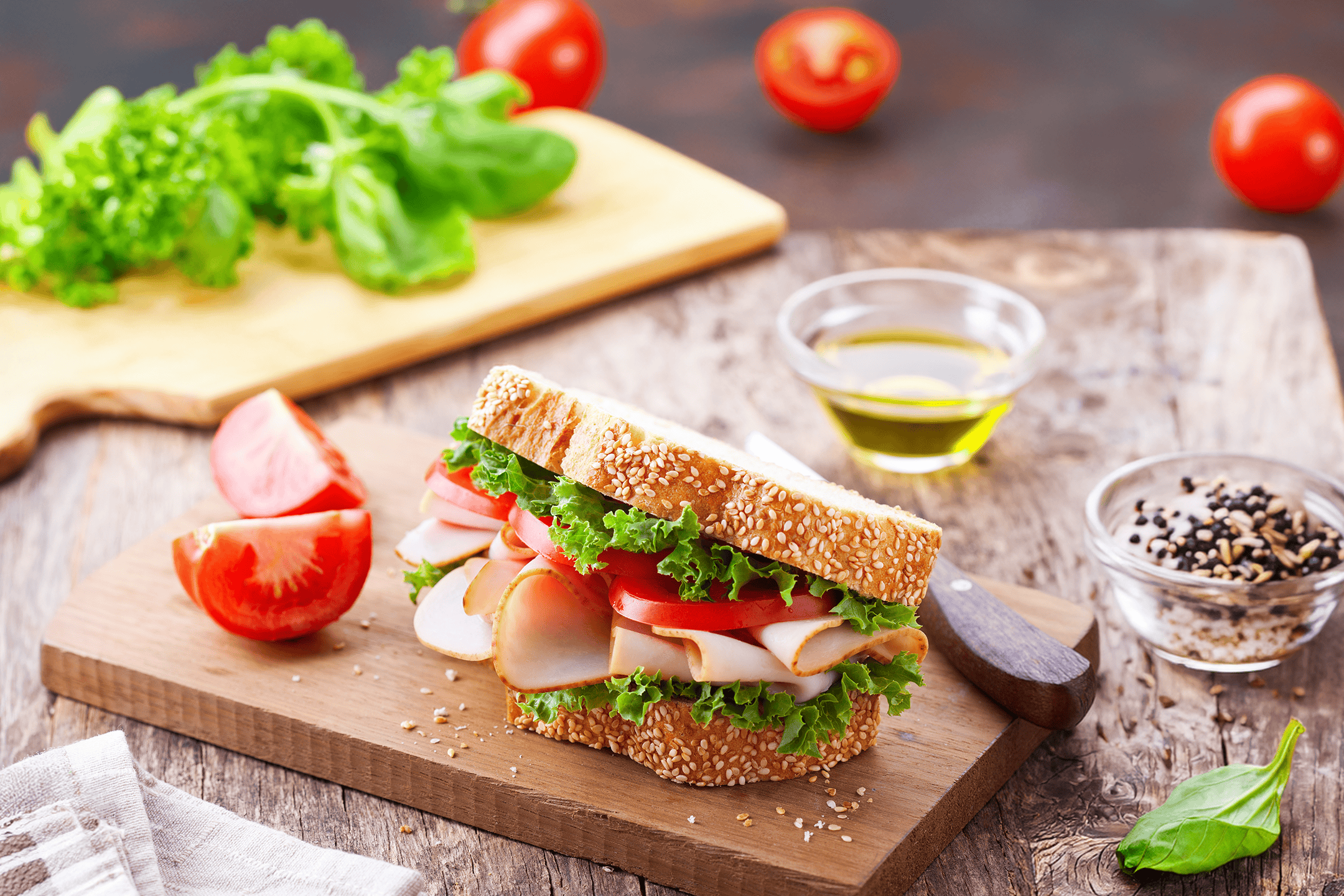 En sandwich med kalkun, salat og tomat på et skærebræt med friske grøntsager.