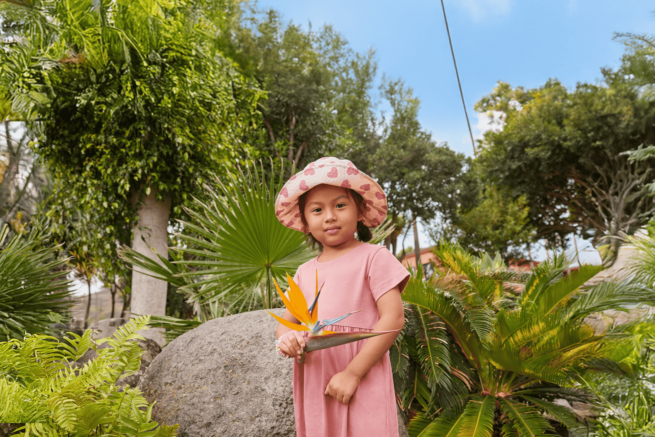 Lille pige i lyserød kjole og hat holder en paradisfuglblomst i en frodig have.