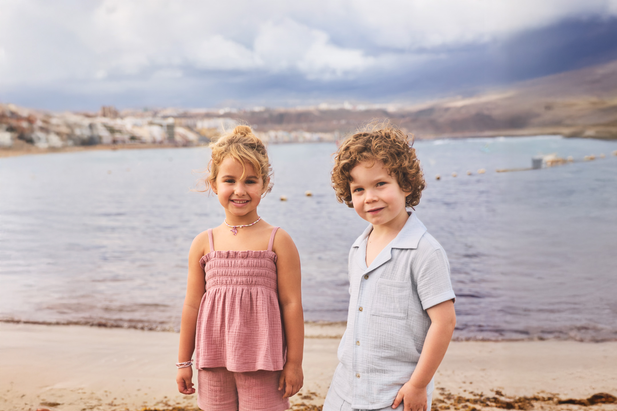 To børn på stranden iført sommeroutfits, en pige i lyserød og en dreng i lyseblå.