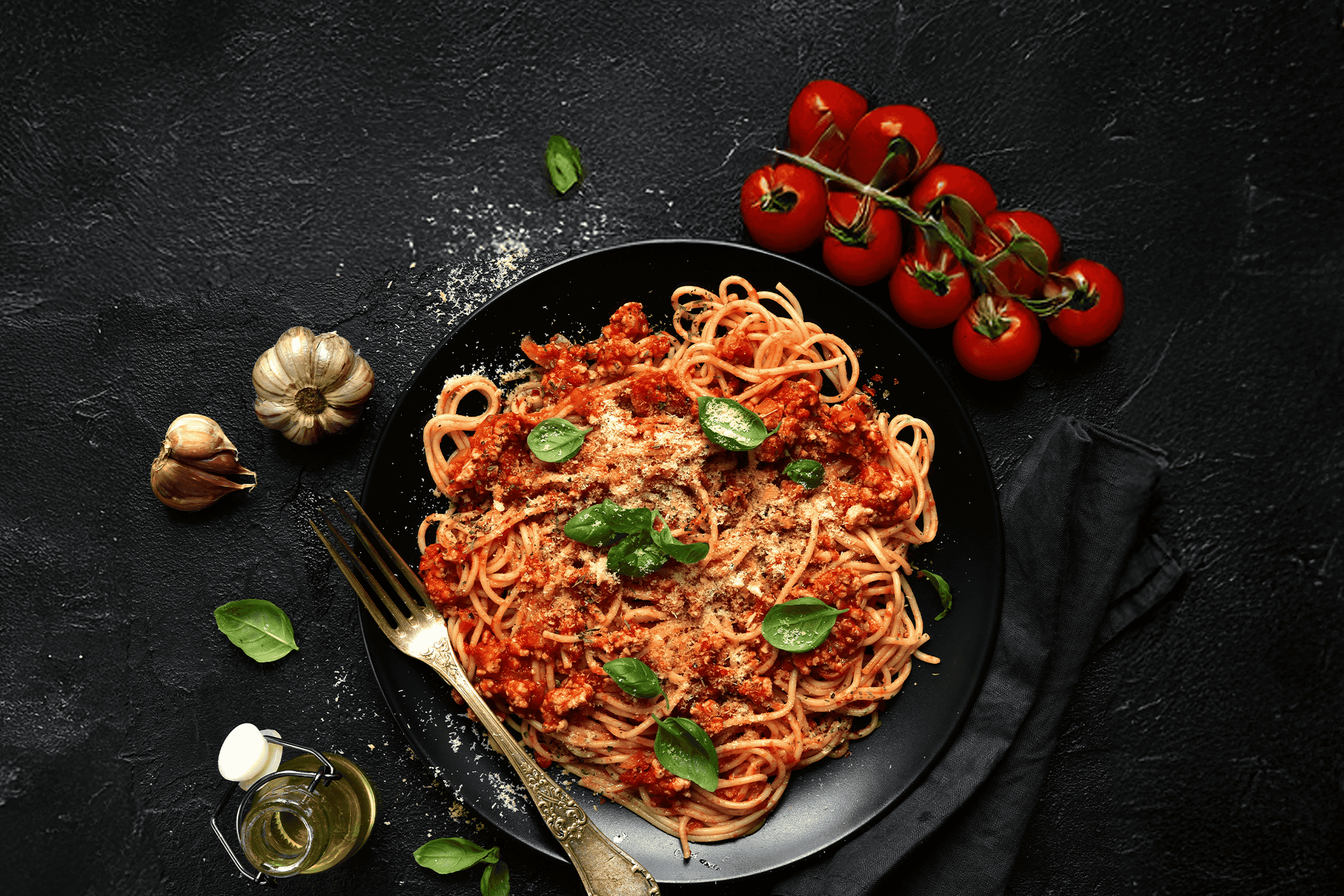 Spaghetti bolognese med basilikum, parmesan, hvidløg, tomater og olivenolie på sort baggrund.