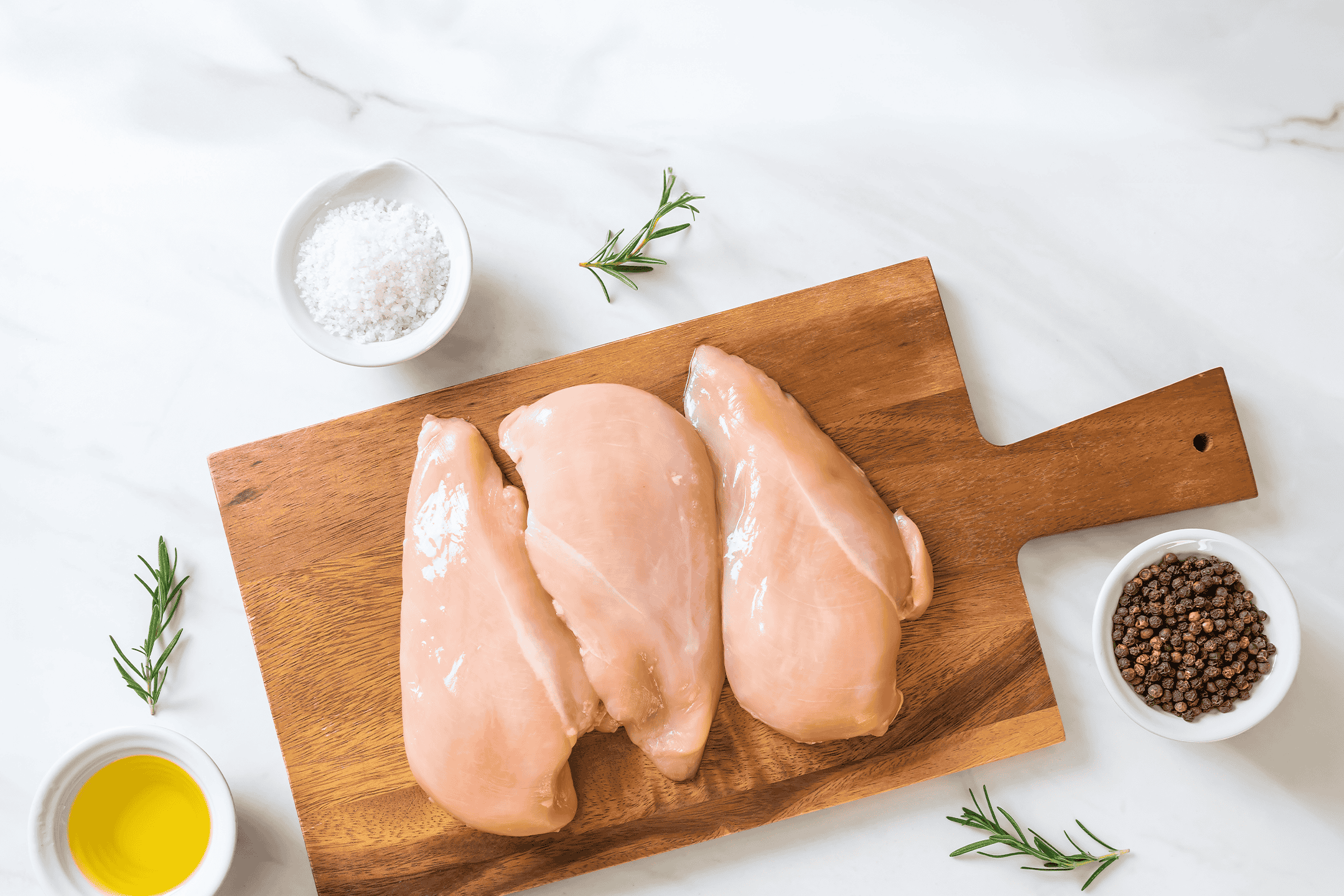 Rå kyllingebrystfileter på et skærebræt med salt, peberkorn, rosmarin og olivenolie.