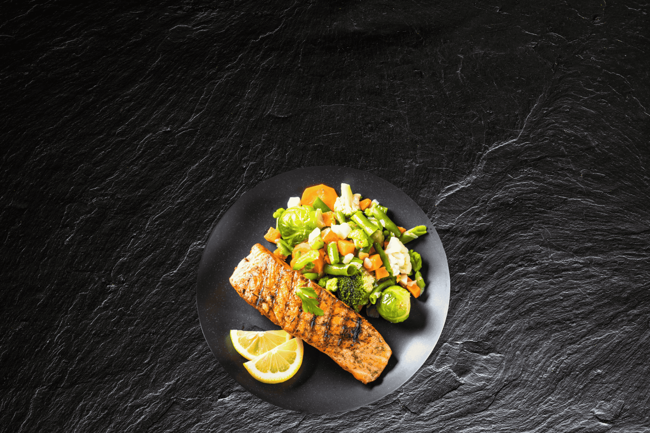 Grillet laks med dampede grøntsager og citron på en sort tallerken.
