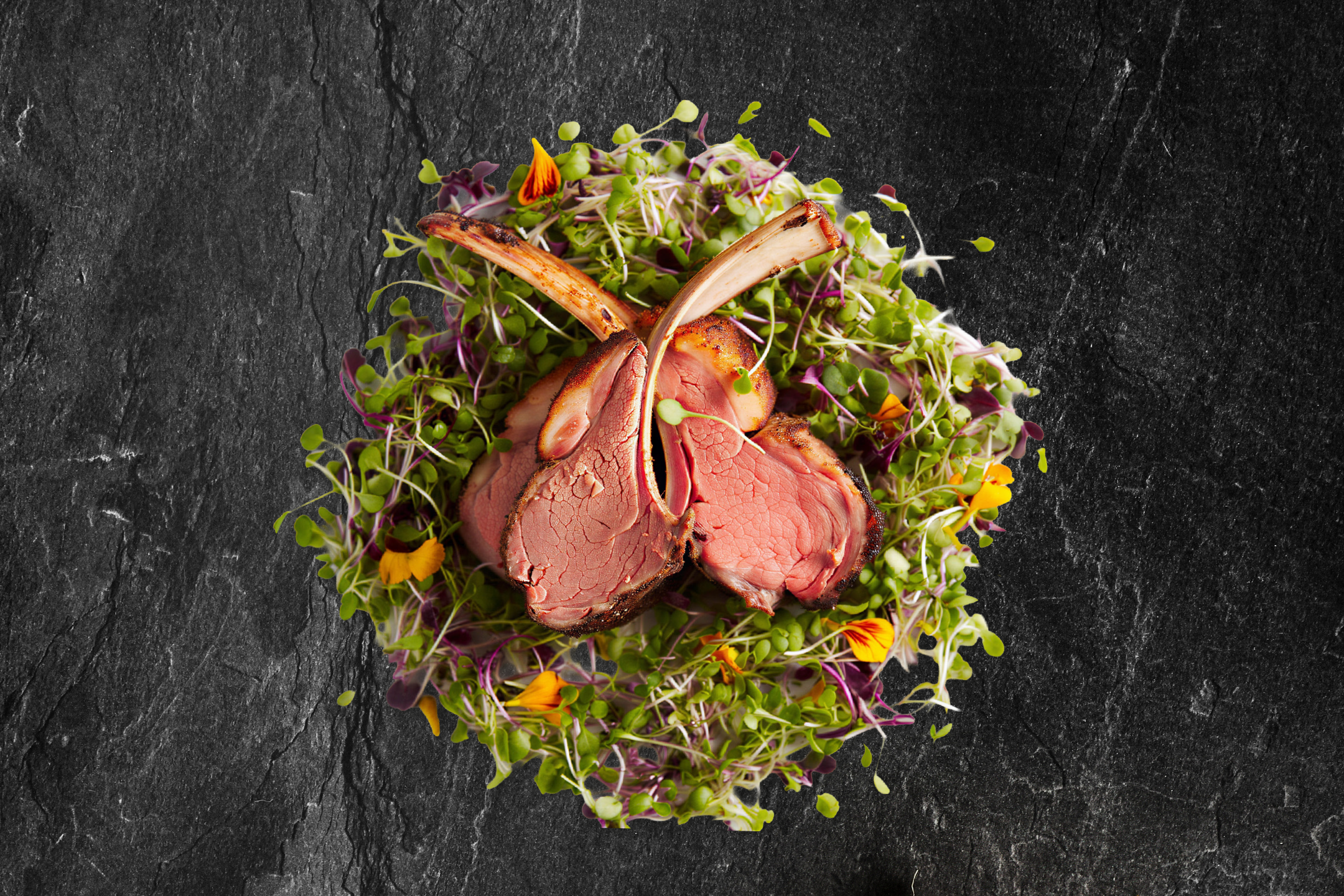 Lammekoteletter med friske spirer og spiselige blomster på en mørk skiferplade.