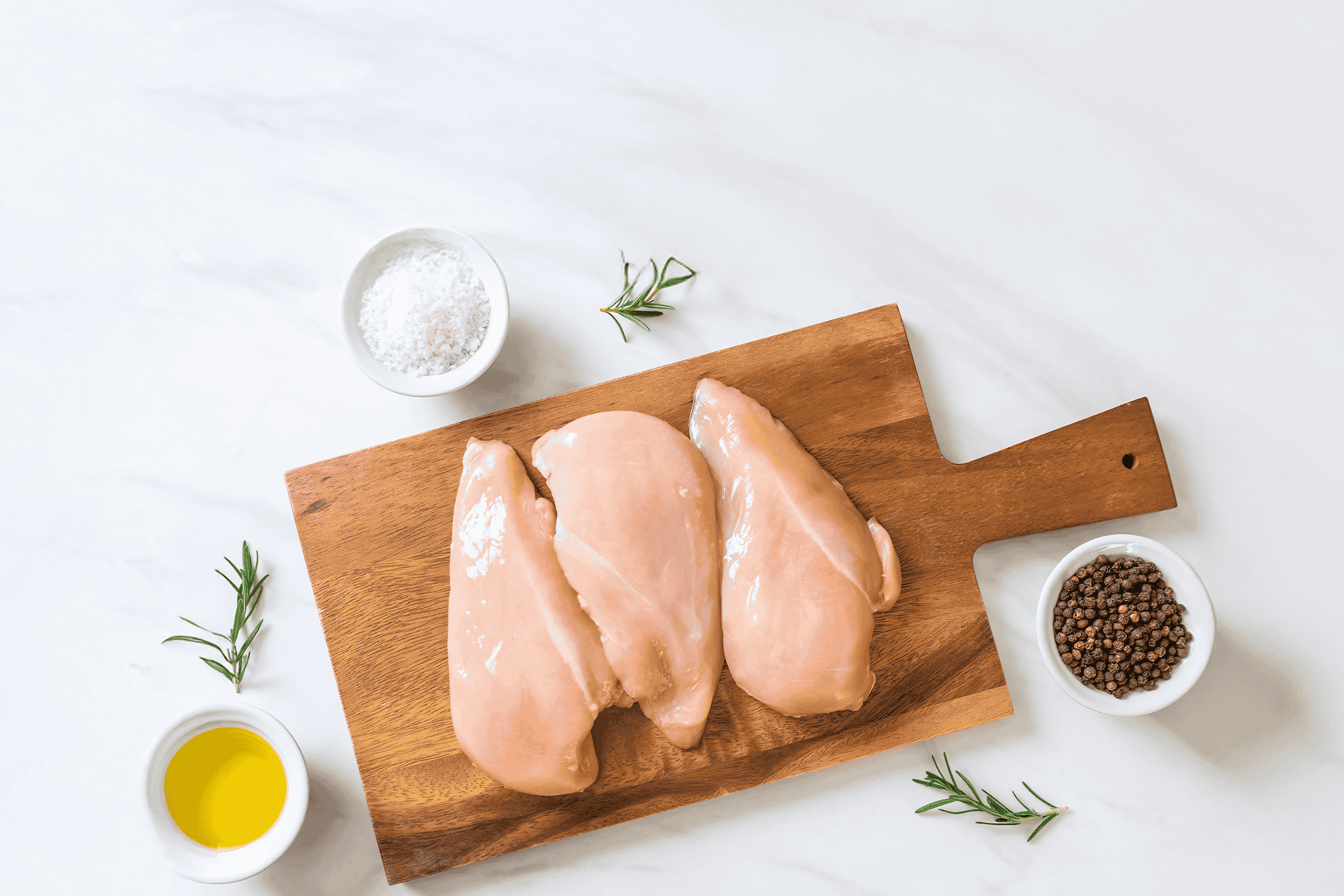 Rå kyllingebrystfileter på et skærebræt med rosmarin, salt, peber og olivenolie.