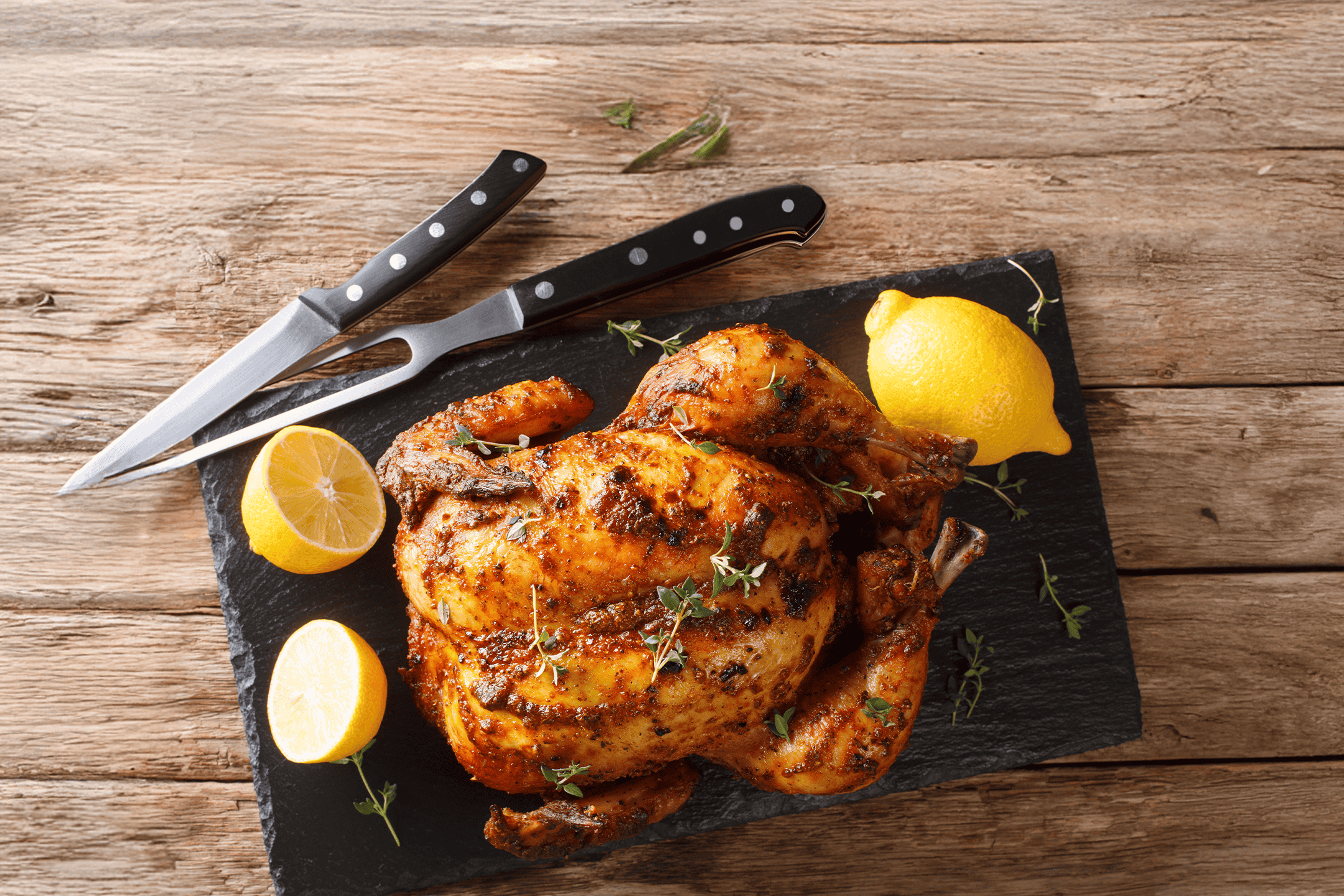 Helstegt kylling med citron og timian på et sort skærebræt med udskæringsbestik.