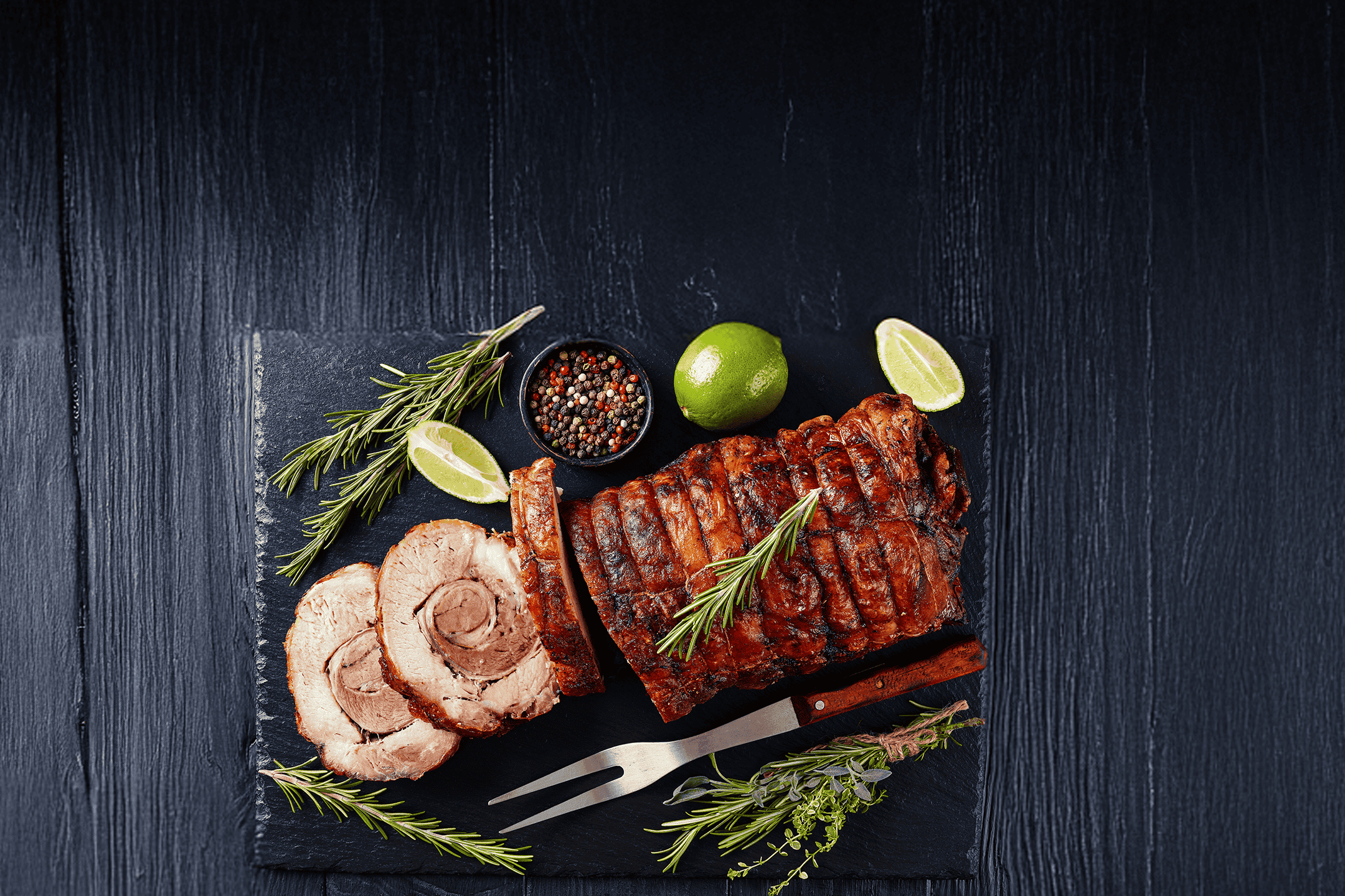 Skiver af grillet svinekød med rosmarin, lime og peberkorn på en sort skiferplade.