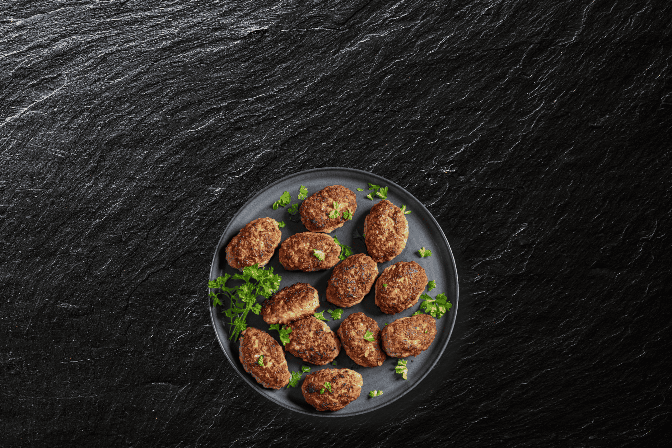 Frikadeller med persille på en mørk tallerken, set ovenfra på en sort skiferbaggrund.