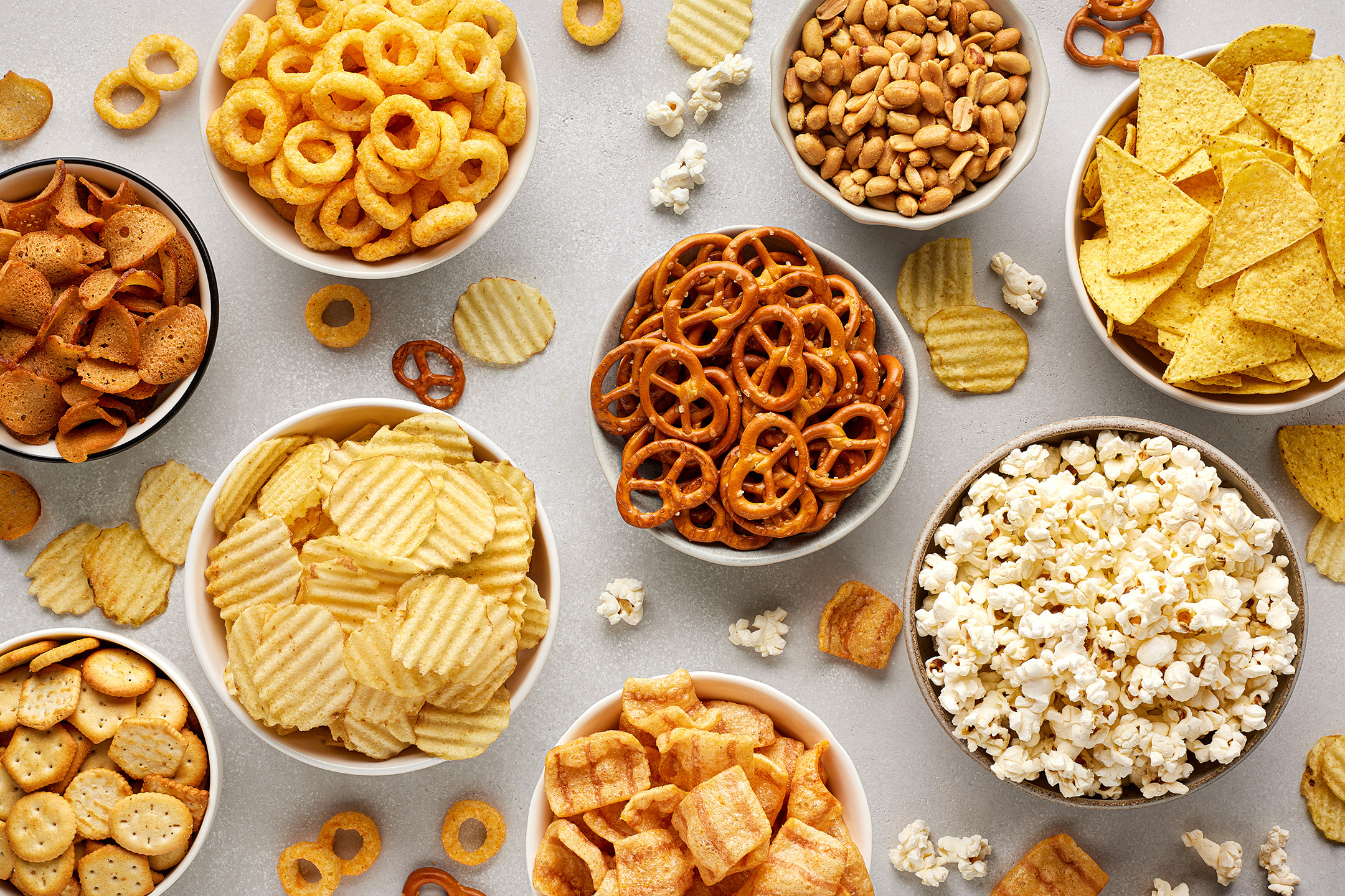 En række skåle fyldt med forskellige snacks som chips, popcorn, peanuts og pretzels.