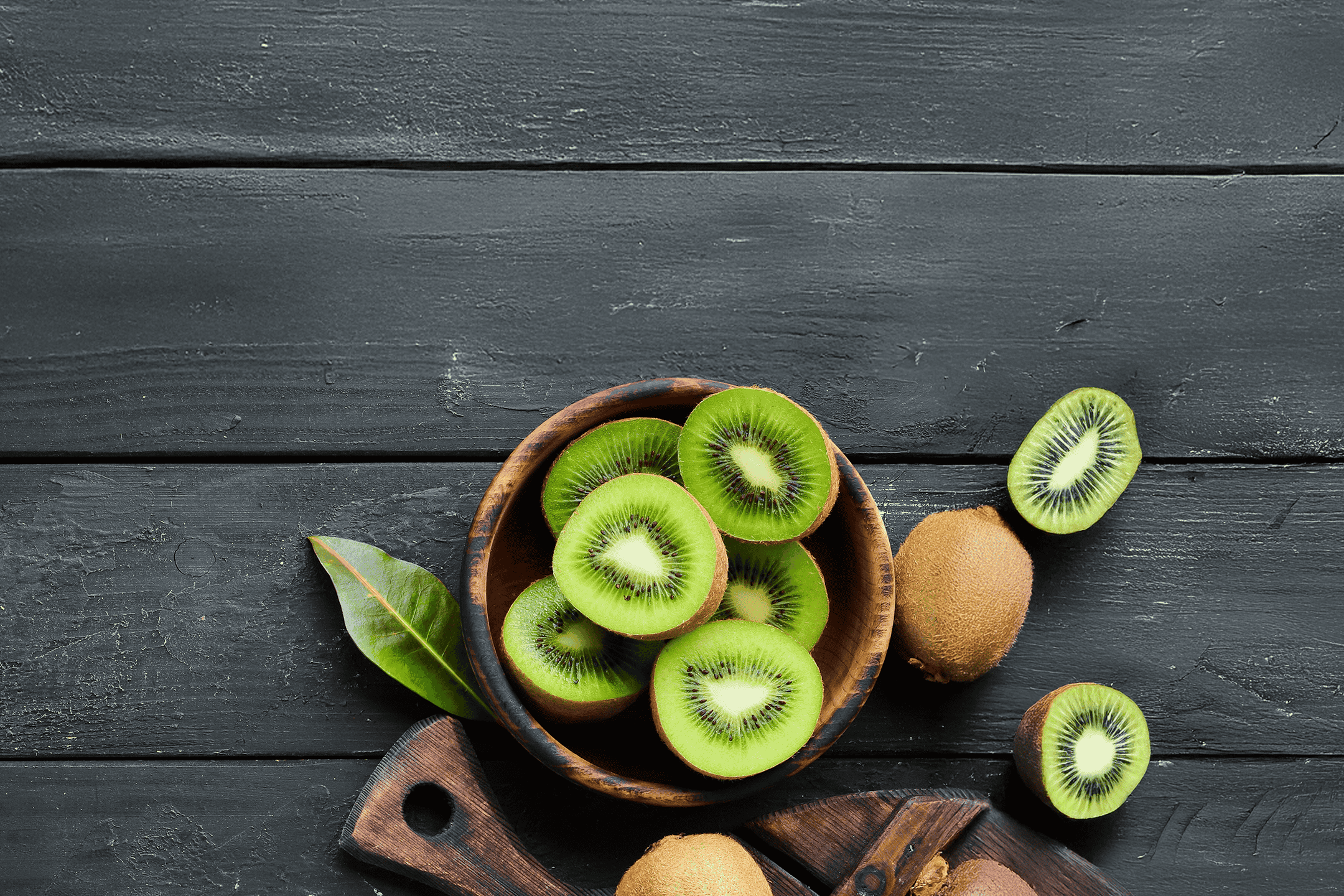 Skåret kiwi i en træskål og hele kiwier på et mørkt træbord.