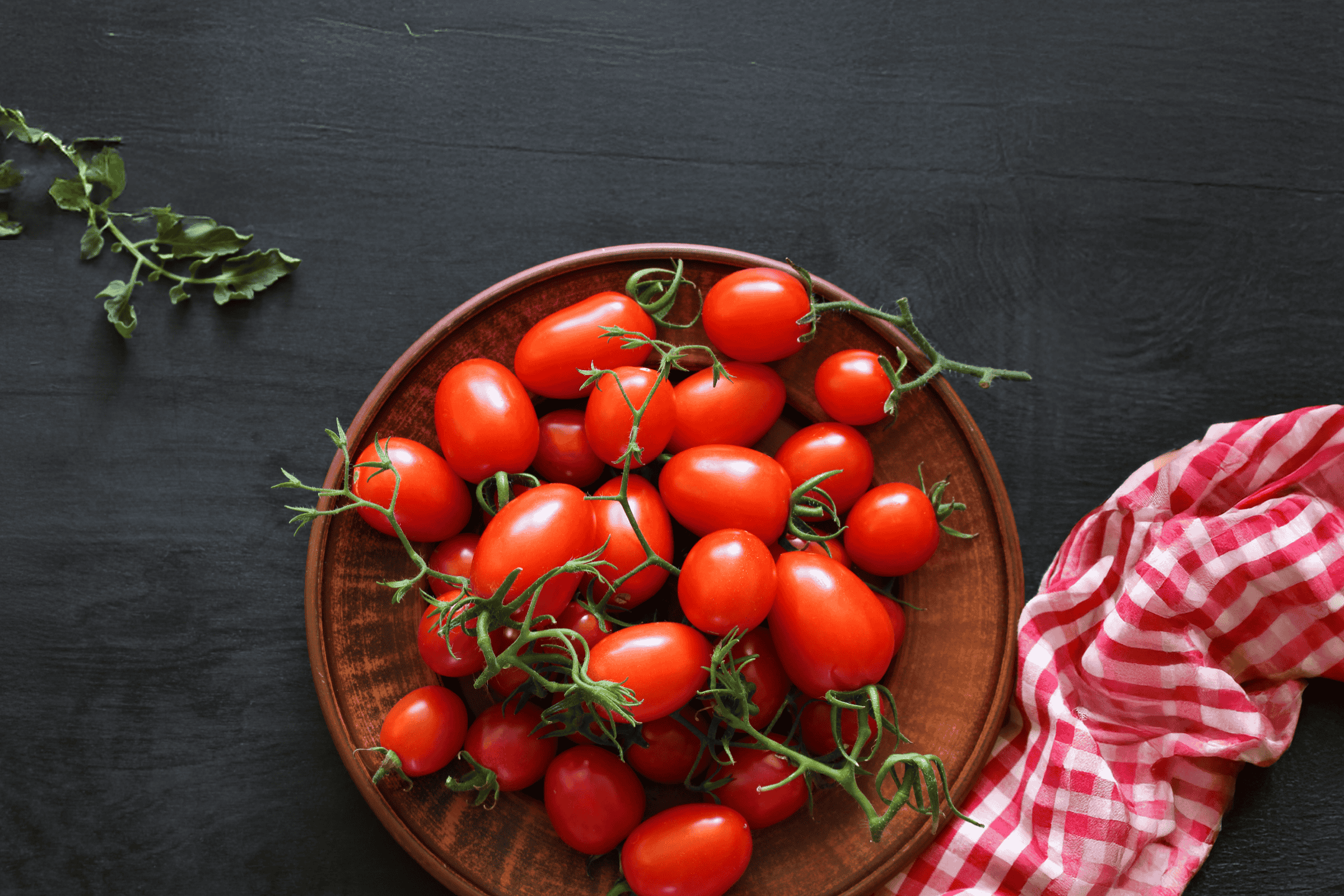 Friske røde cherrytomater i en brun skål på et mørkt træbord med et rødt og hvidt viskestykke.