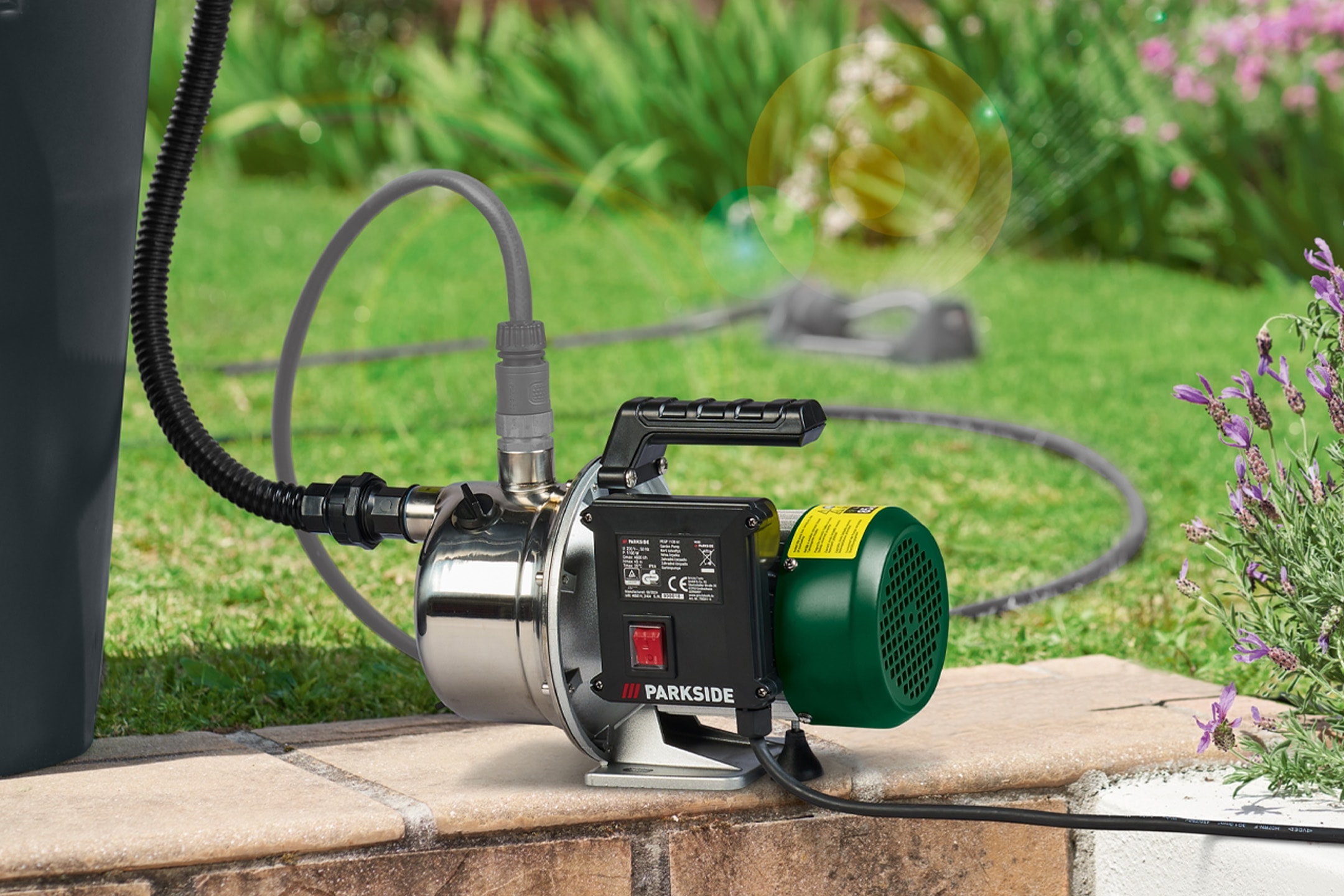 Vandpumpe med slanger og ledninger tilsluttet en regnvandstønde, der vander en have.