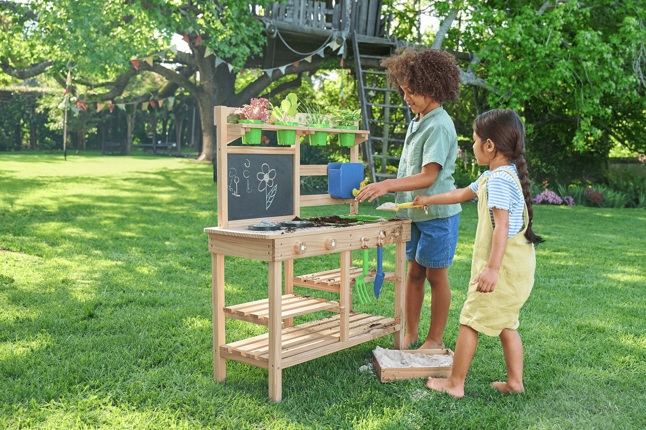 To børn leger med et udendørs legekøkken med jord og planter i en have.