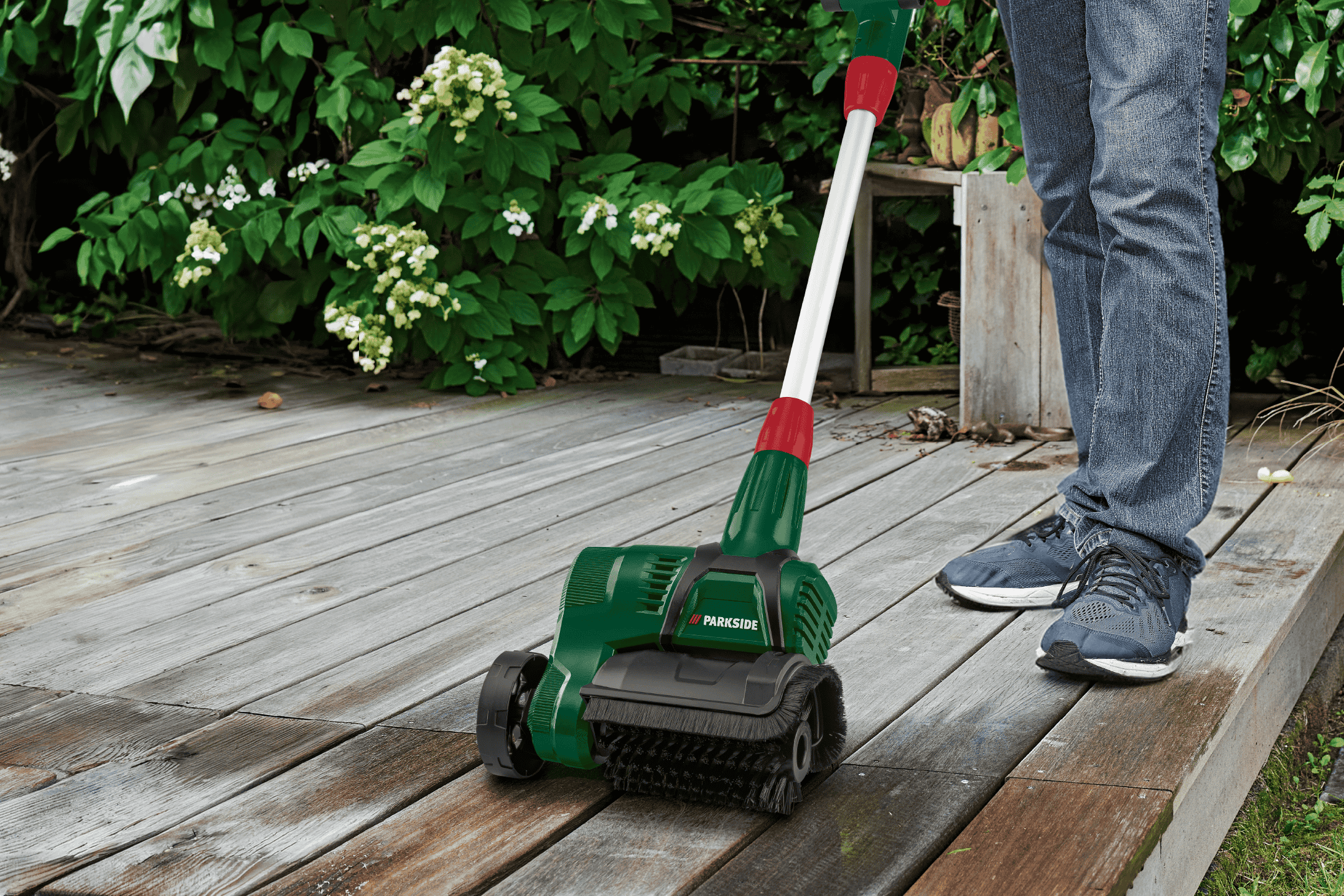 En person rengør et trædæk med en elektrisk terrassebørste, omgivet af grønne planter.