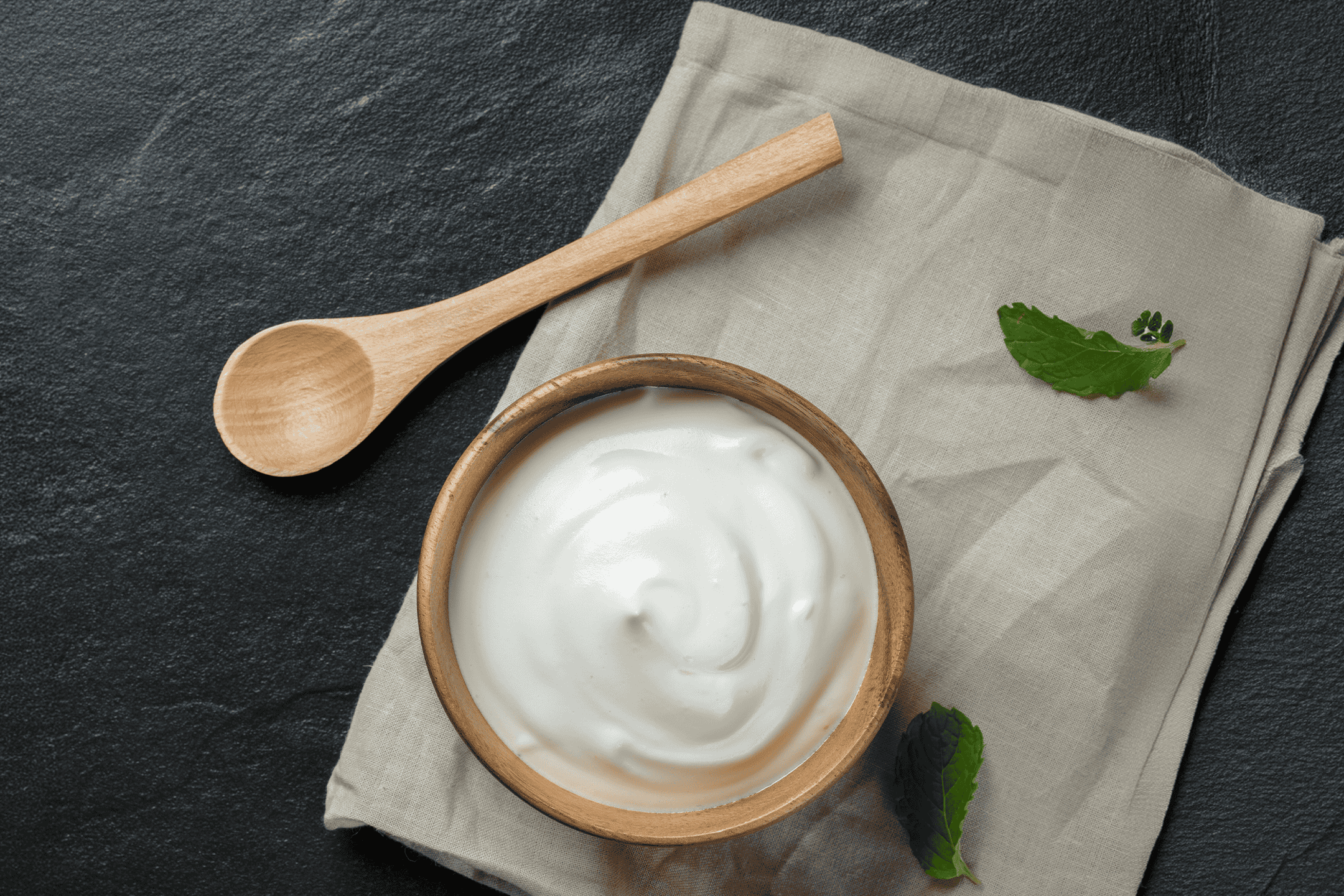 En skål med yoghurt, en træske og et viskestykke på en mørk baggrund.