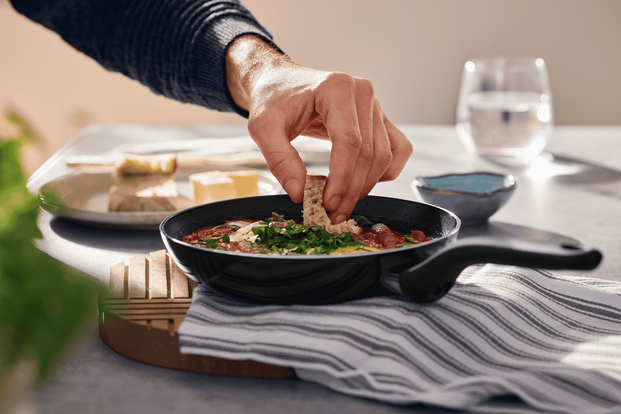 En person dypper brød i en pande med shakshuka, med et glas vand og ost i baggrunden.