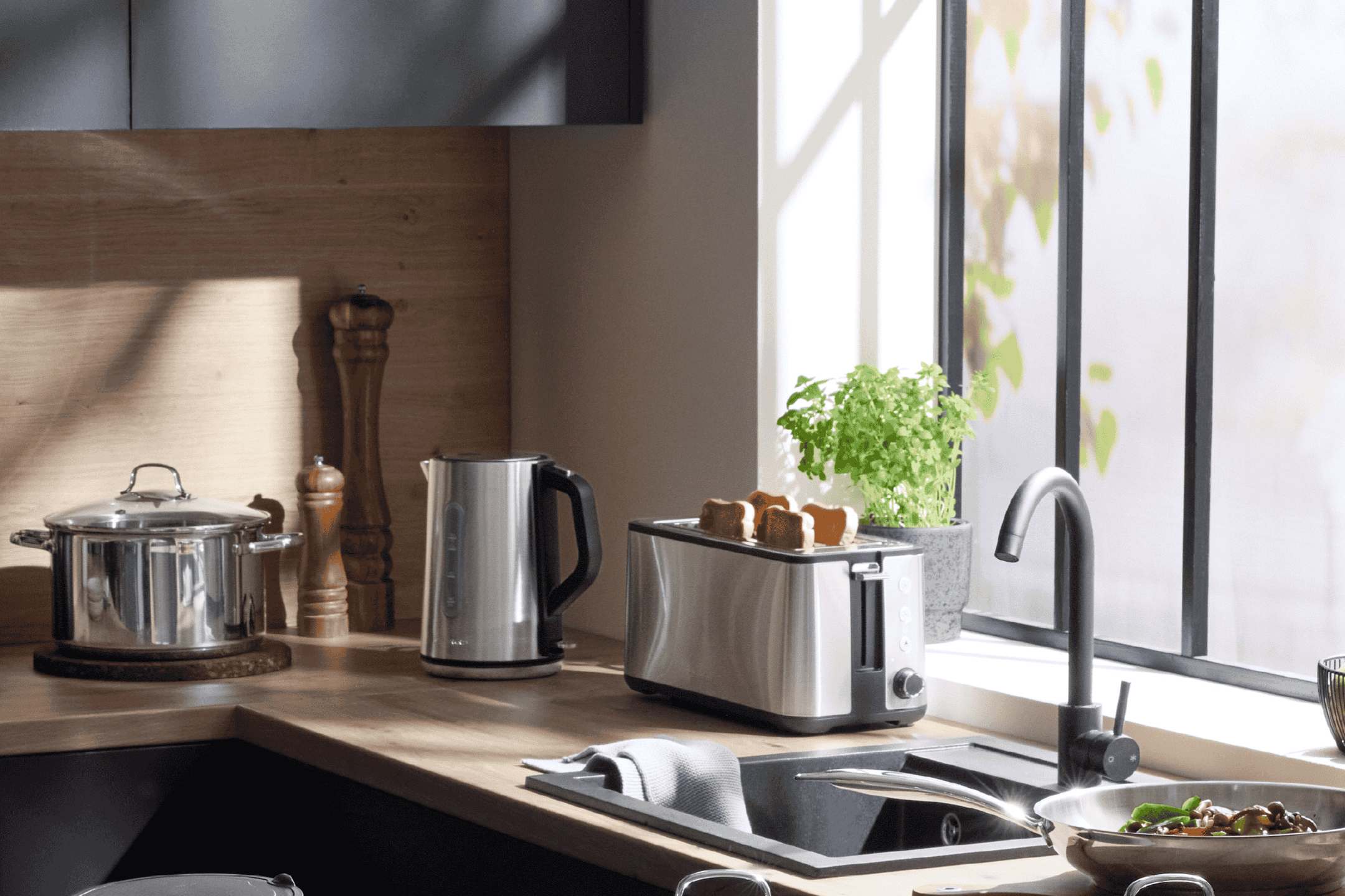 Køkkenbord med køkkenudstyr i rustfrit stål, herunder en elkedel, brødrister og gryde.