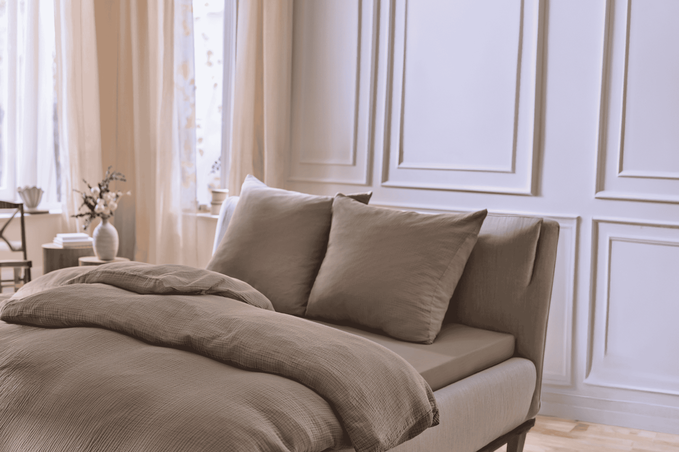 Soveværelse med seng med brunt sengetøj og puder, hvide vægge og gardiner.