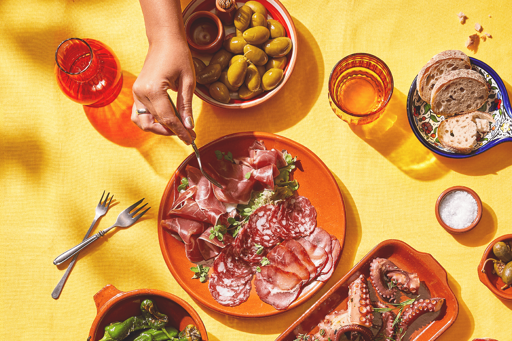 En person tager charcuteri fra et fad med oliven, brød, blæksprutte og peberfrugter på et gult bord.